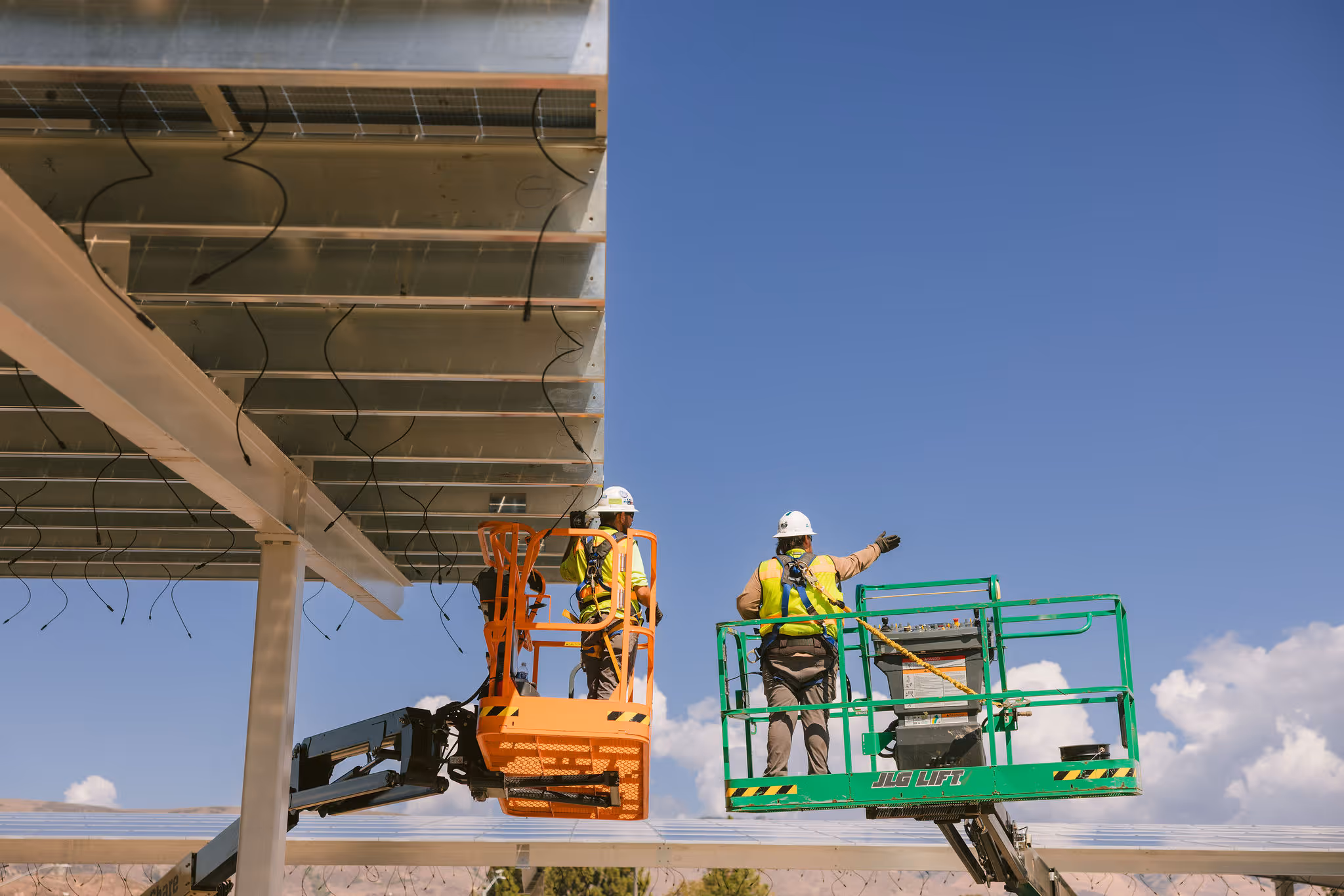 Two construction workers in lift platforms installing solar panels under a clear blue sky.