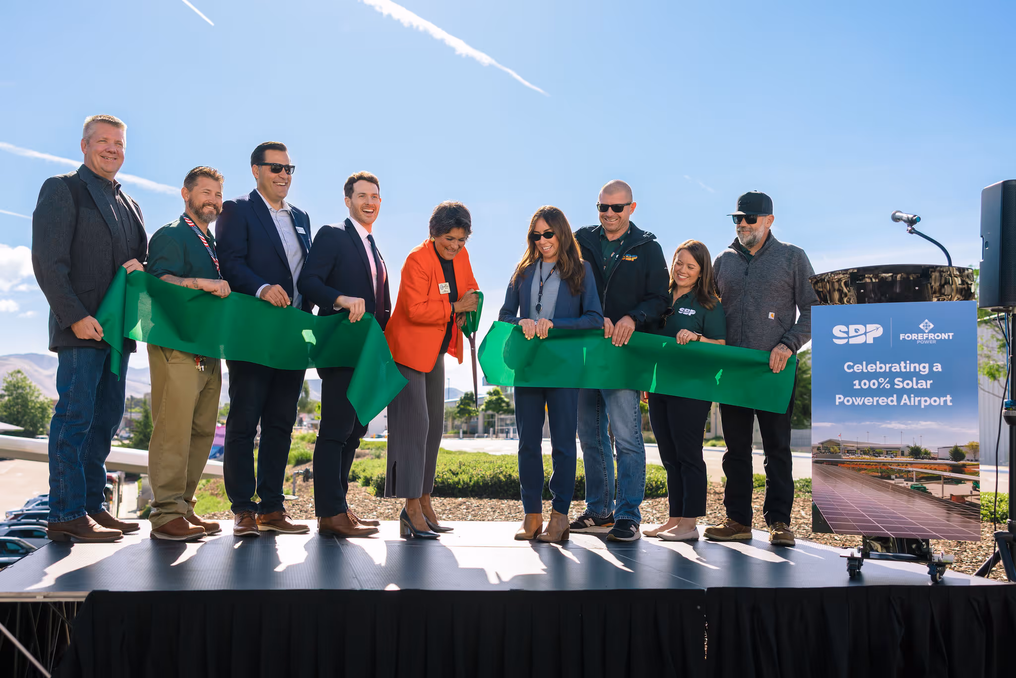 Group cutting a green ribbon at an outdoor event celebrating a 100% solar powered airport.