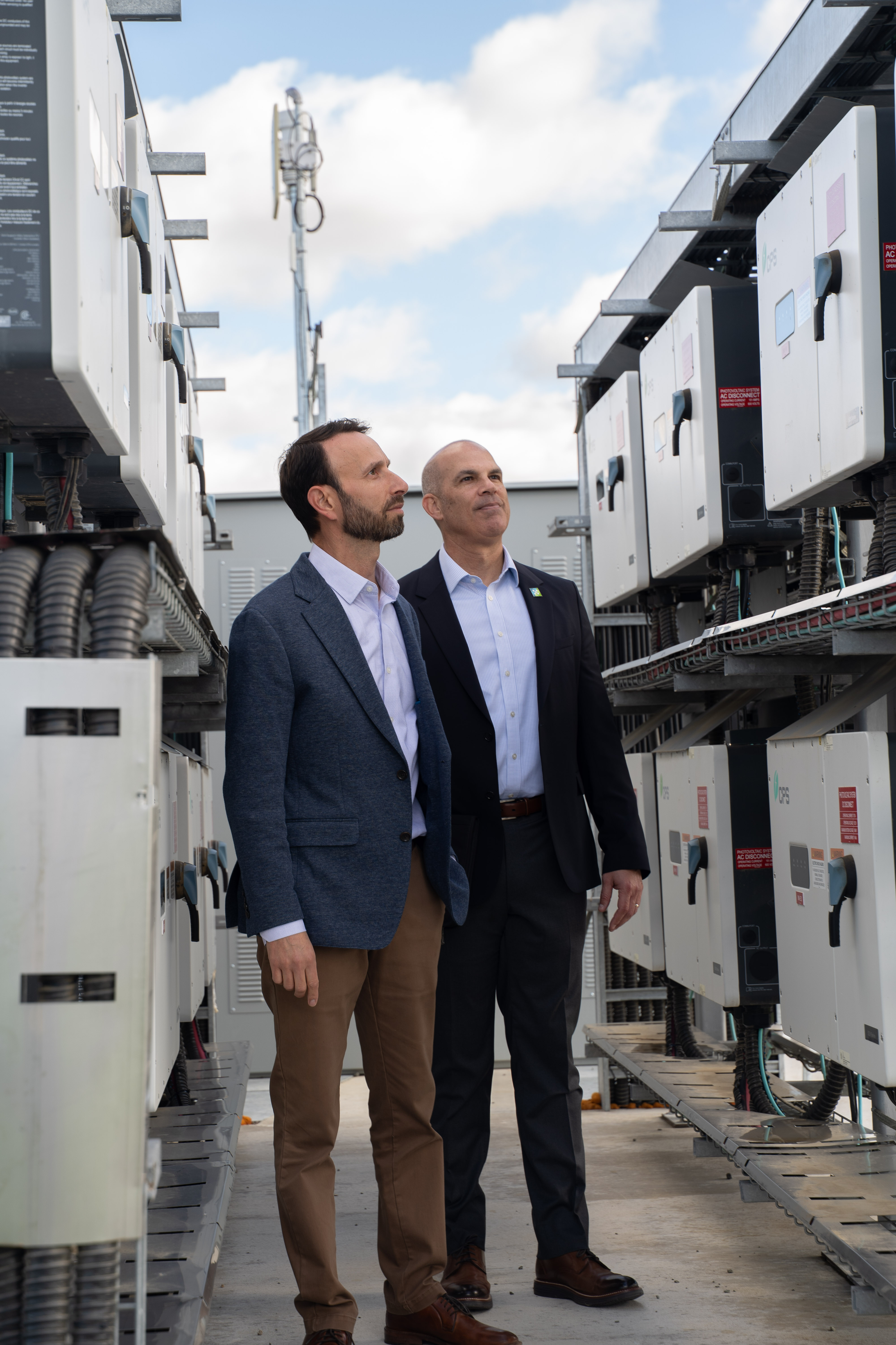 Two men in business attire inspecting electrical equipment outdoors under a partly cloudy sky.