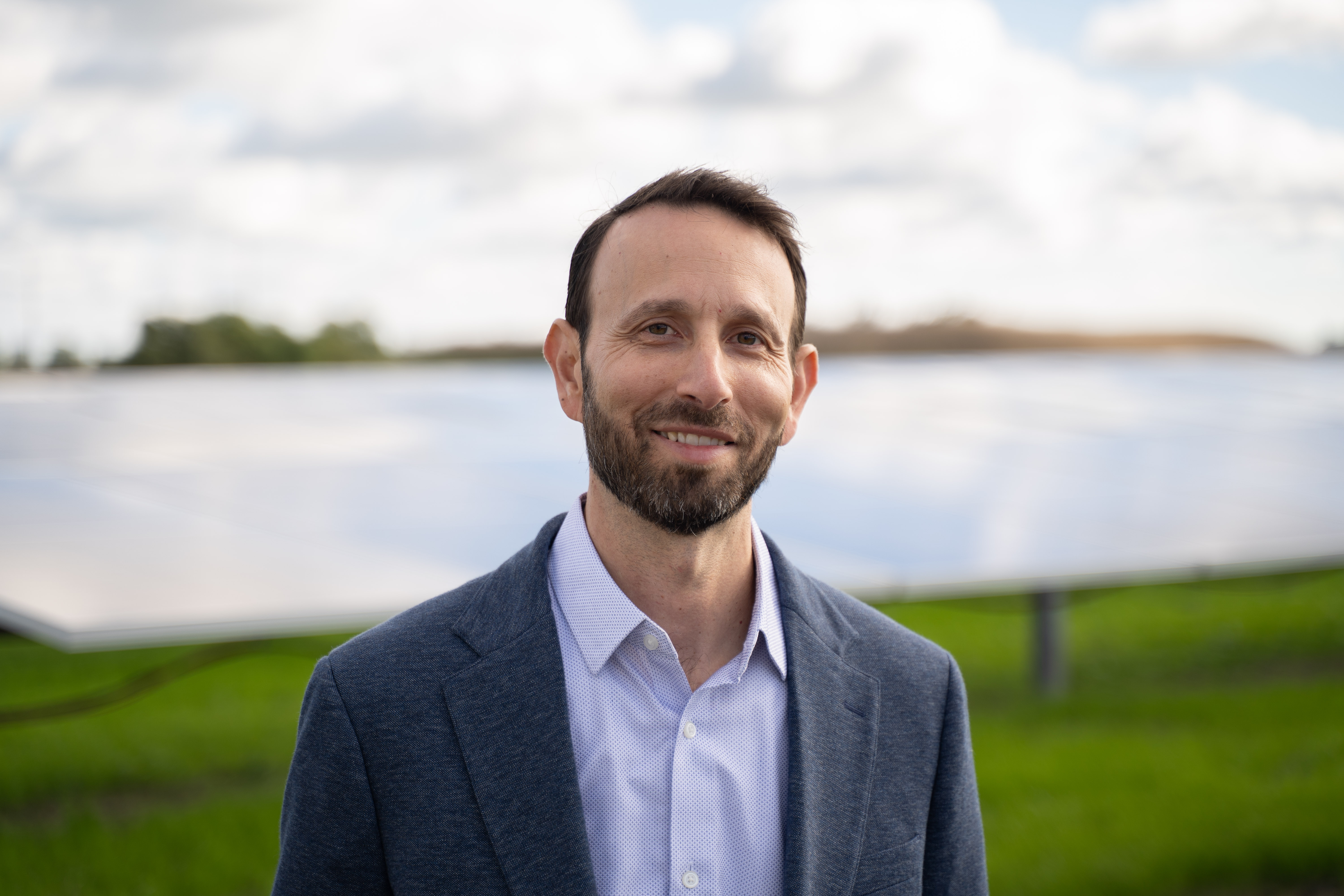 Man with beard wearing a light blue shirt and dark blazer standing outdoors with solar panels in the background.