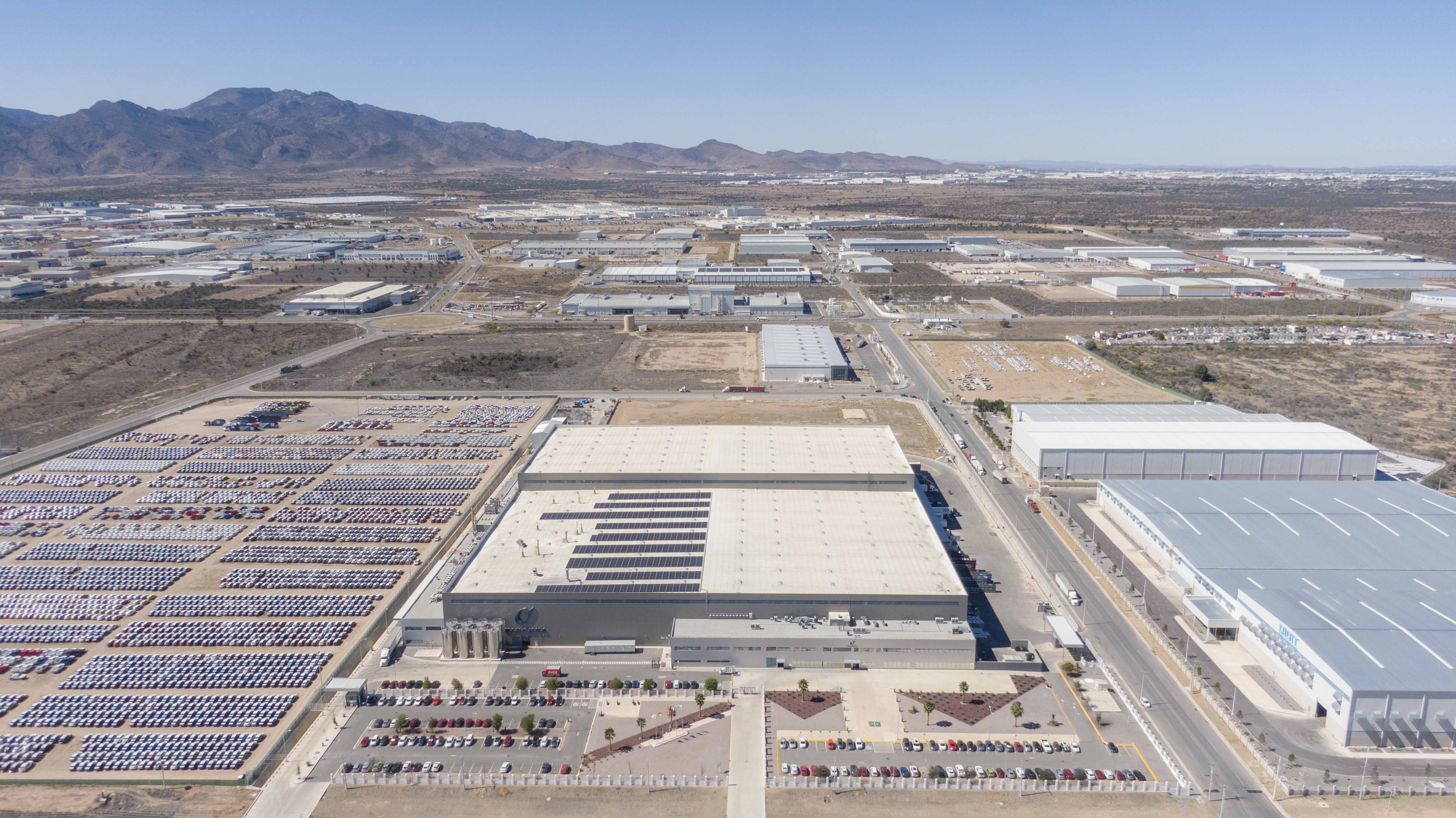 Aerial view of industrial warehouses, parked cars, and mountainous landscape under clear sky.