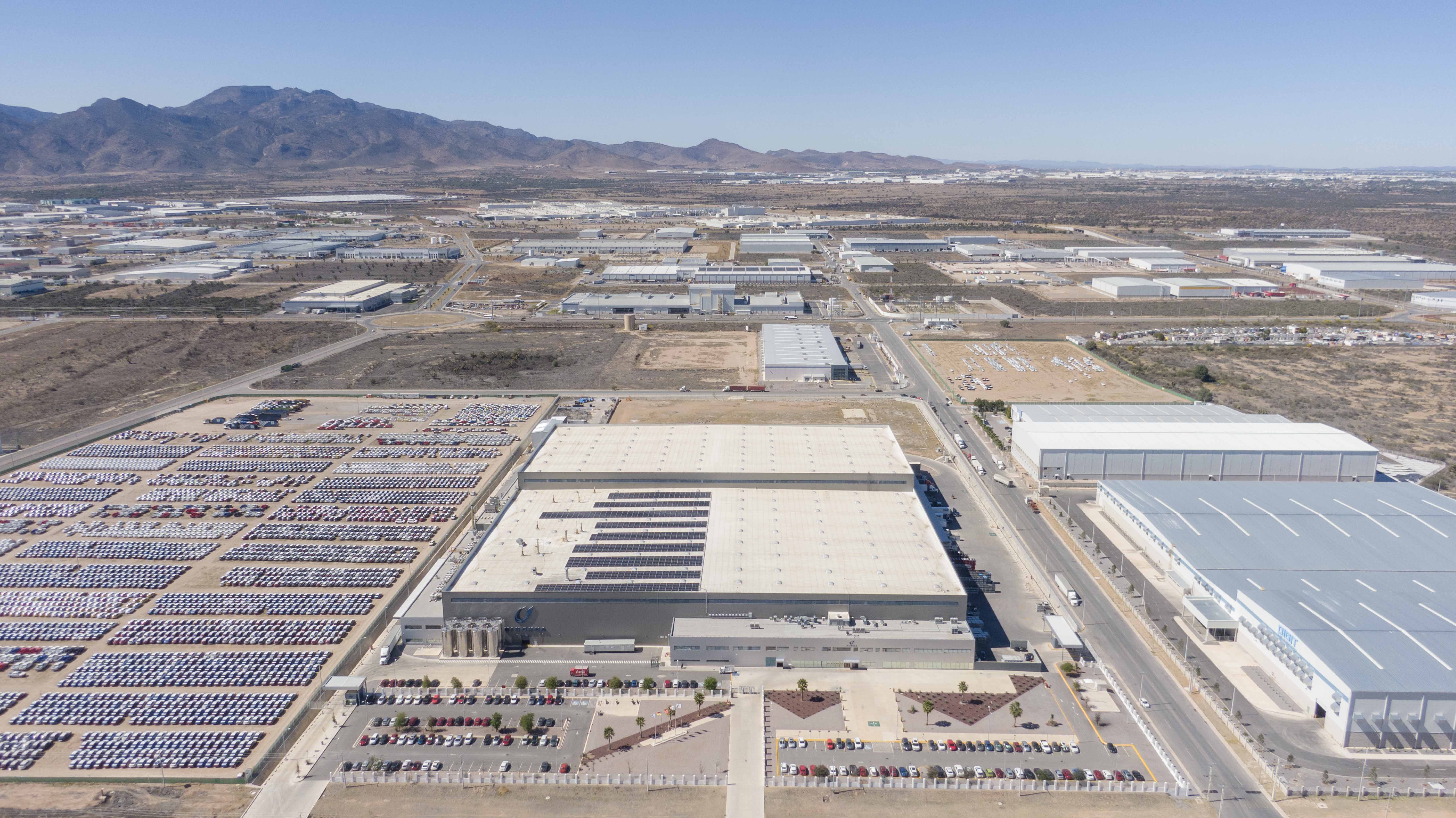 Aerial view of industrial warehouses, parked cars, and mountains under clear blue sky.