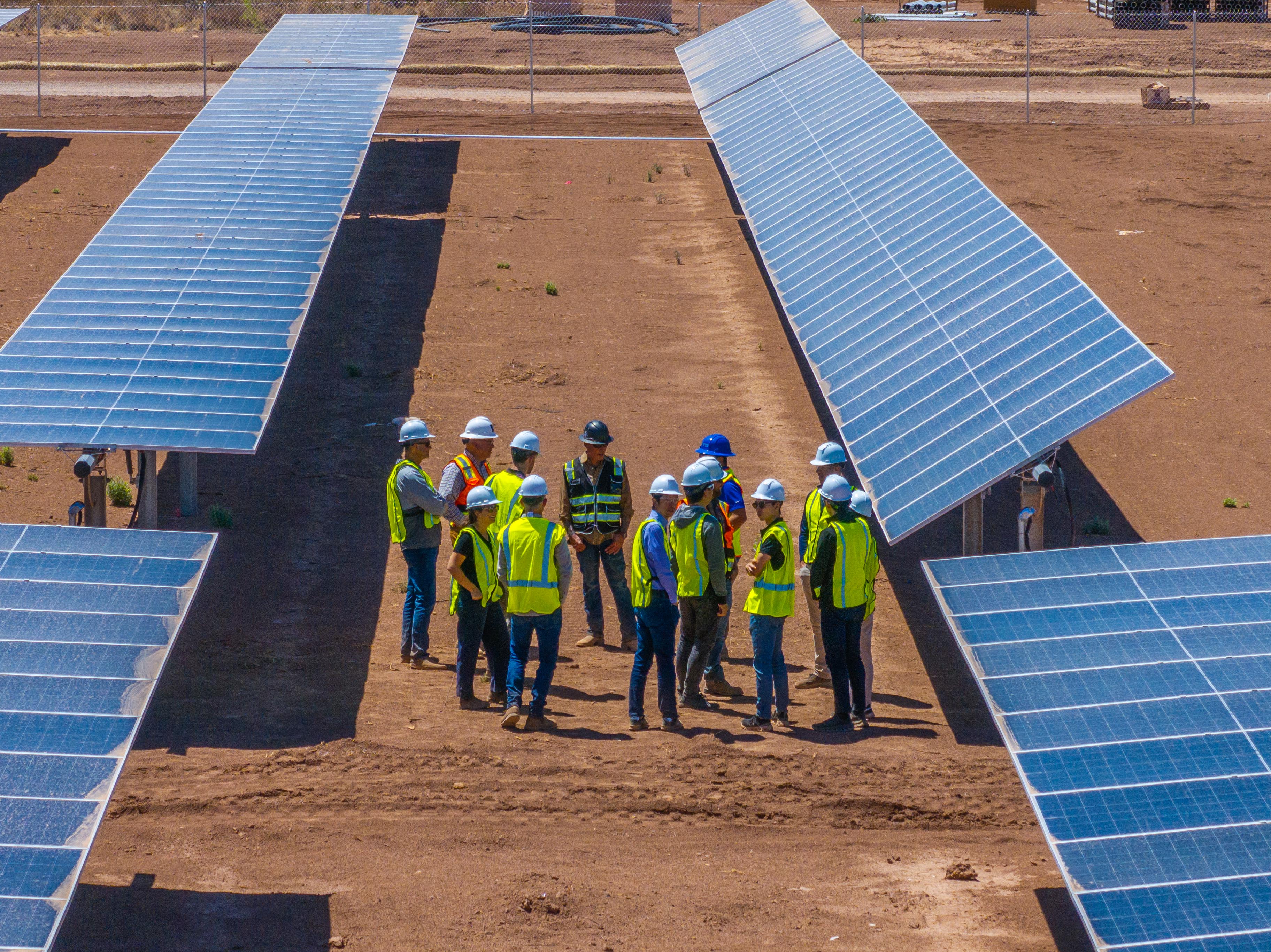 Group of workers in safety vests and helmets standing between solar panel arrays on dry land.