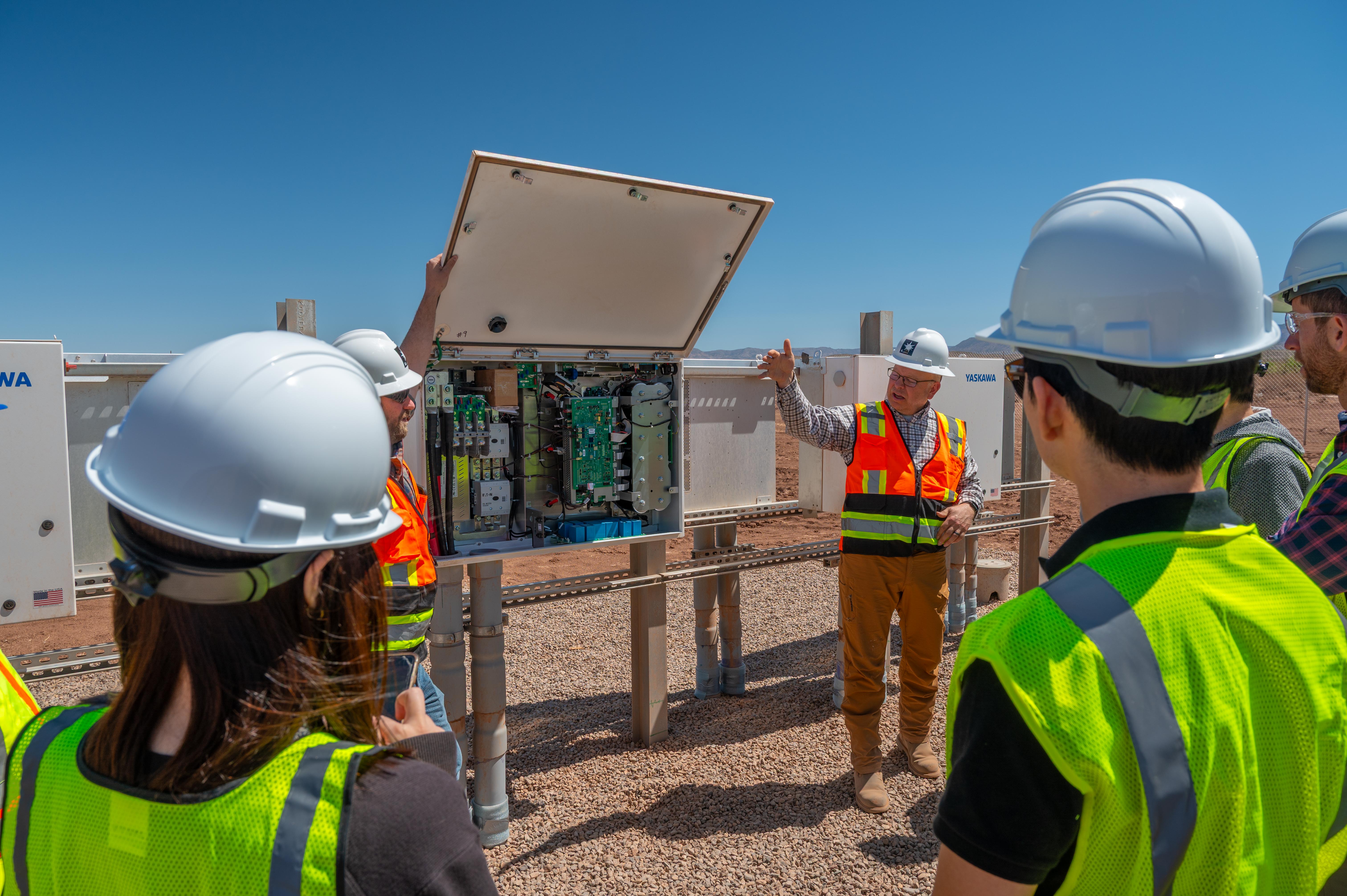 Construction workers in hard hats and safety vests inspecting an open electrical control panel outdoors.