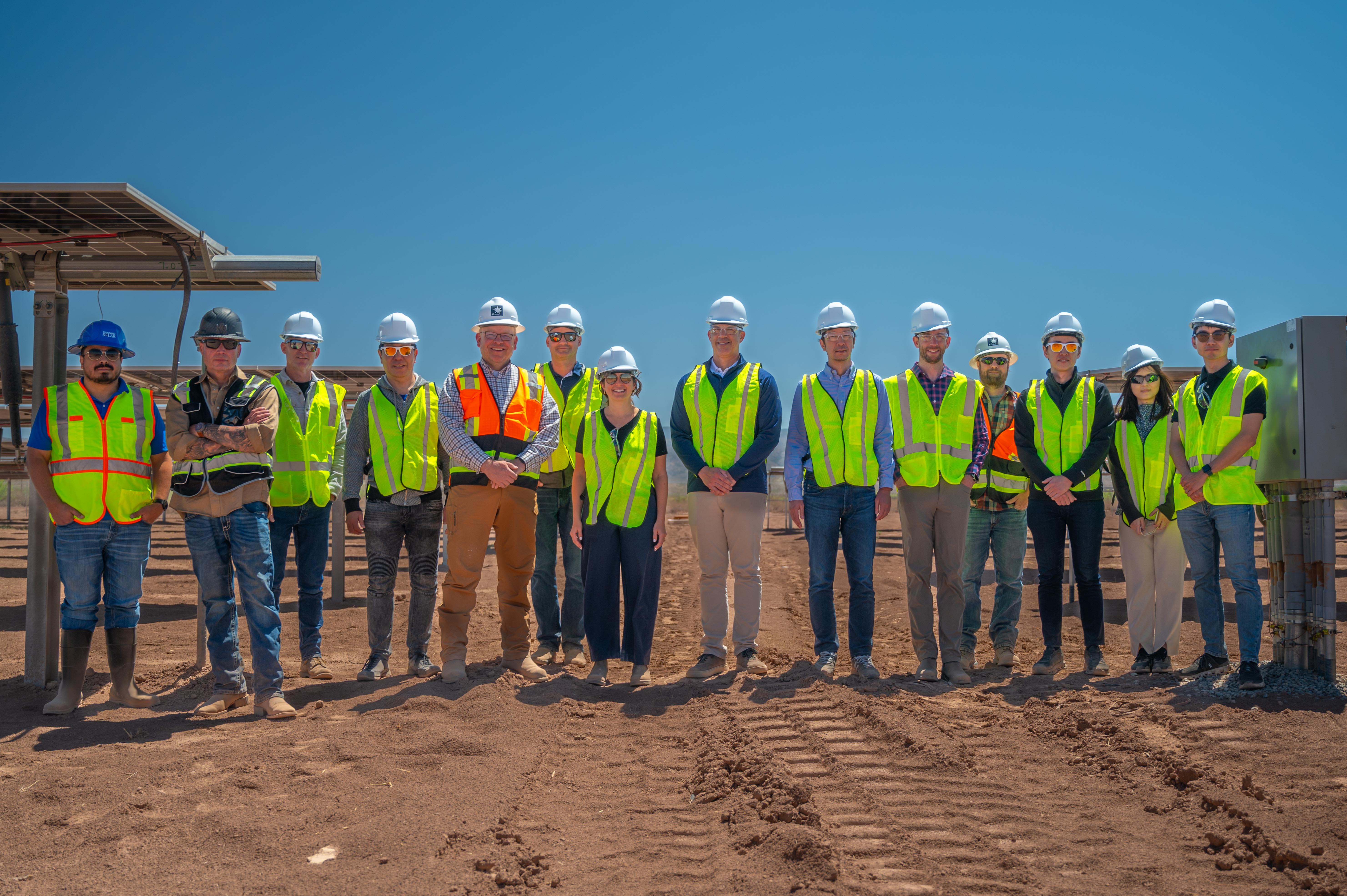 Group of workers wearing safety vests and helmets standing in a solar panel field under a blue sky.