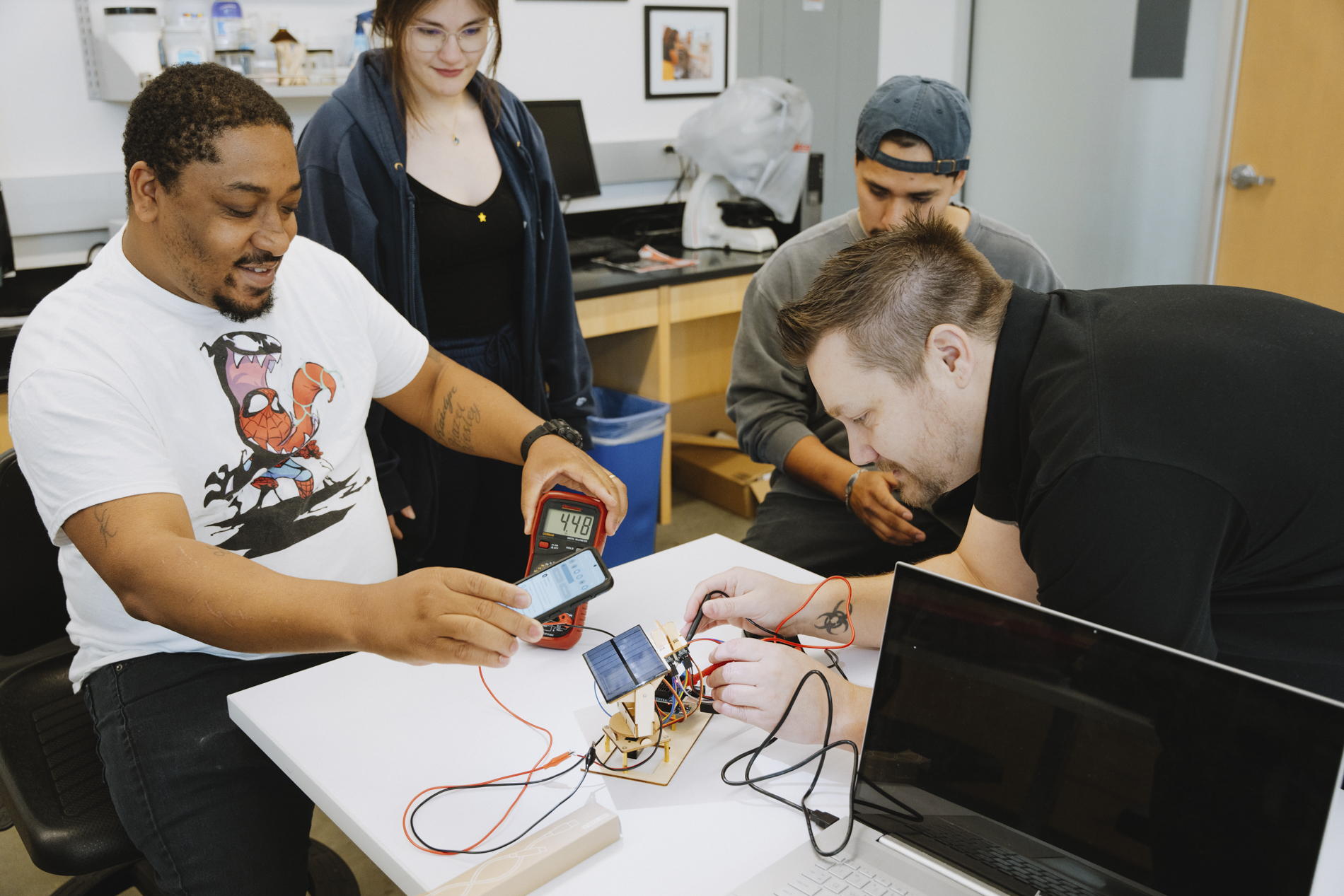 Four people assemble and test a small solar panel device in a tech workspace with laptops and tools.