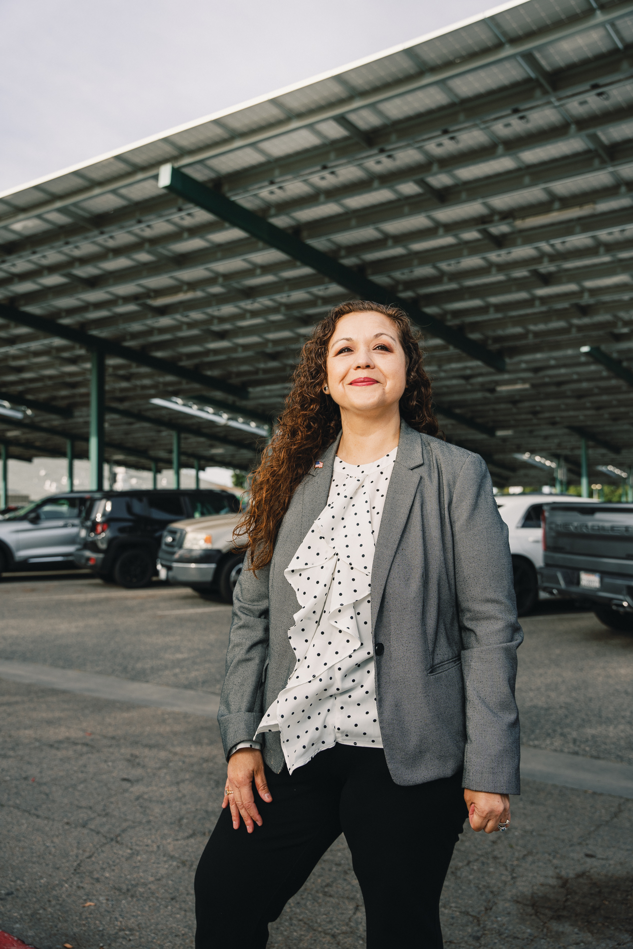 Woman with curly hair wearing a gray blazer and white polka dot blouse standing in a parking lot.