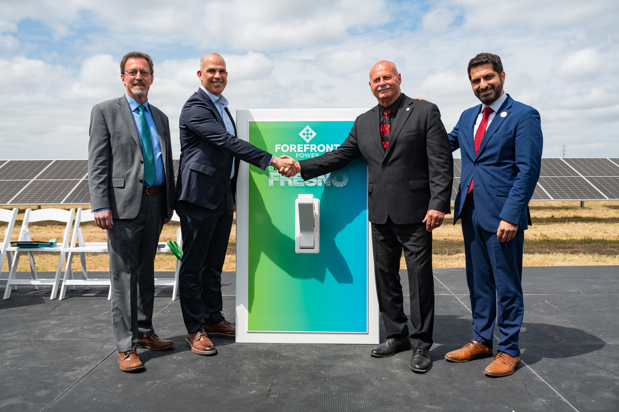 Four men in suits standing outdoors by a large switch panel with solar panels in the background.