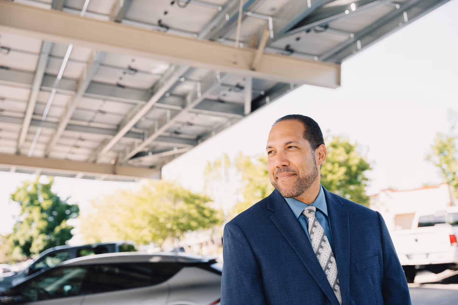Smiling man in blue suit and patterned tie standing outdoors near parked cars under a metal roof.