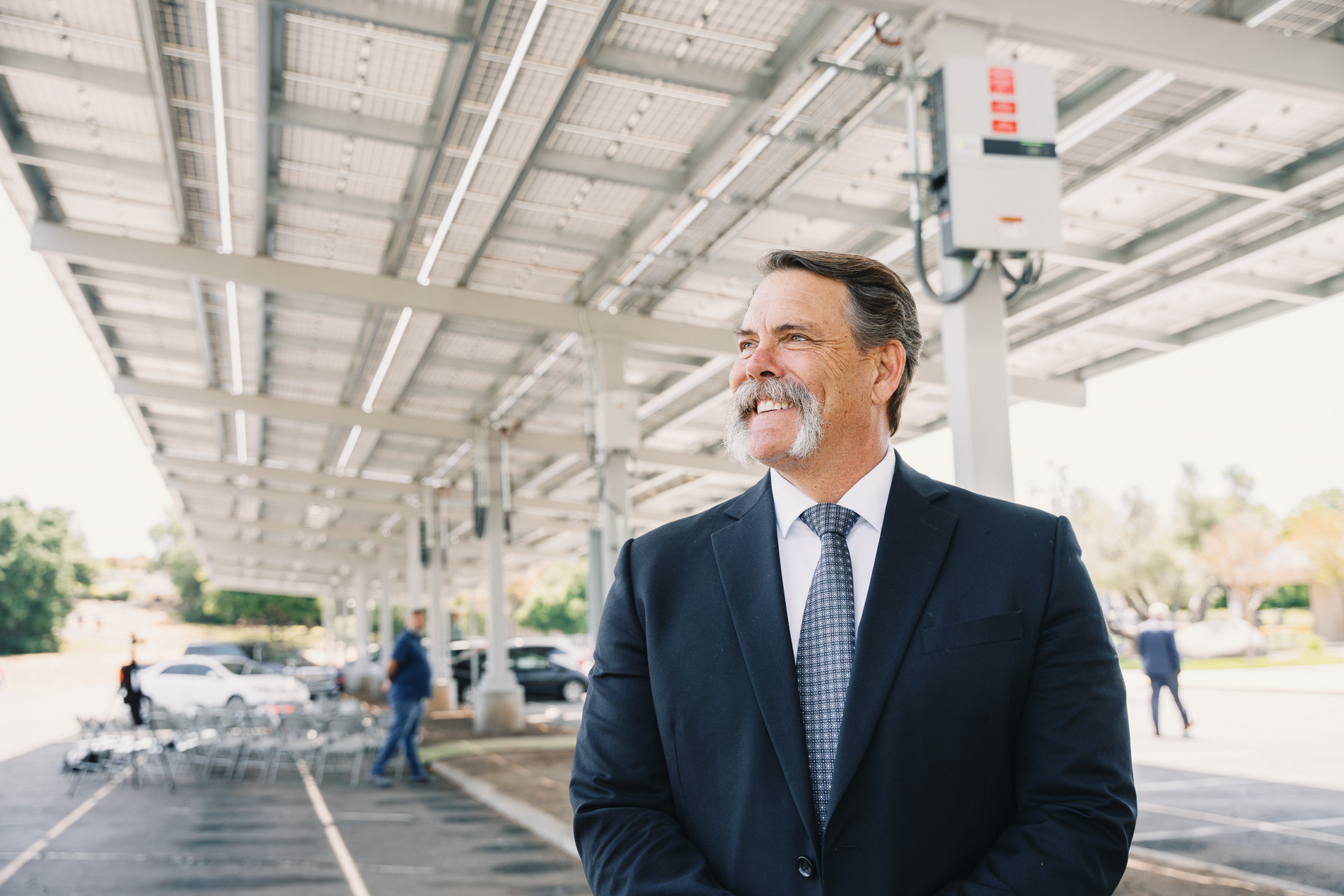 Smiling man in a suit standing under solar panel canopy in parking lot during daytime.