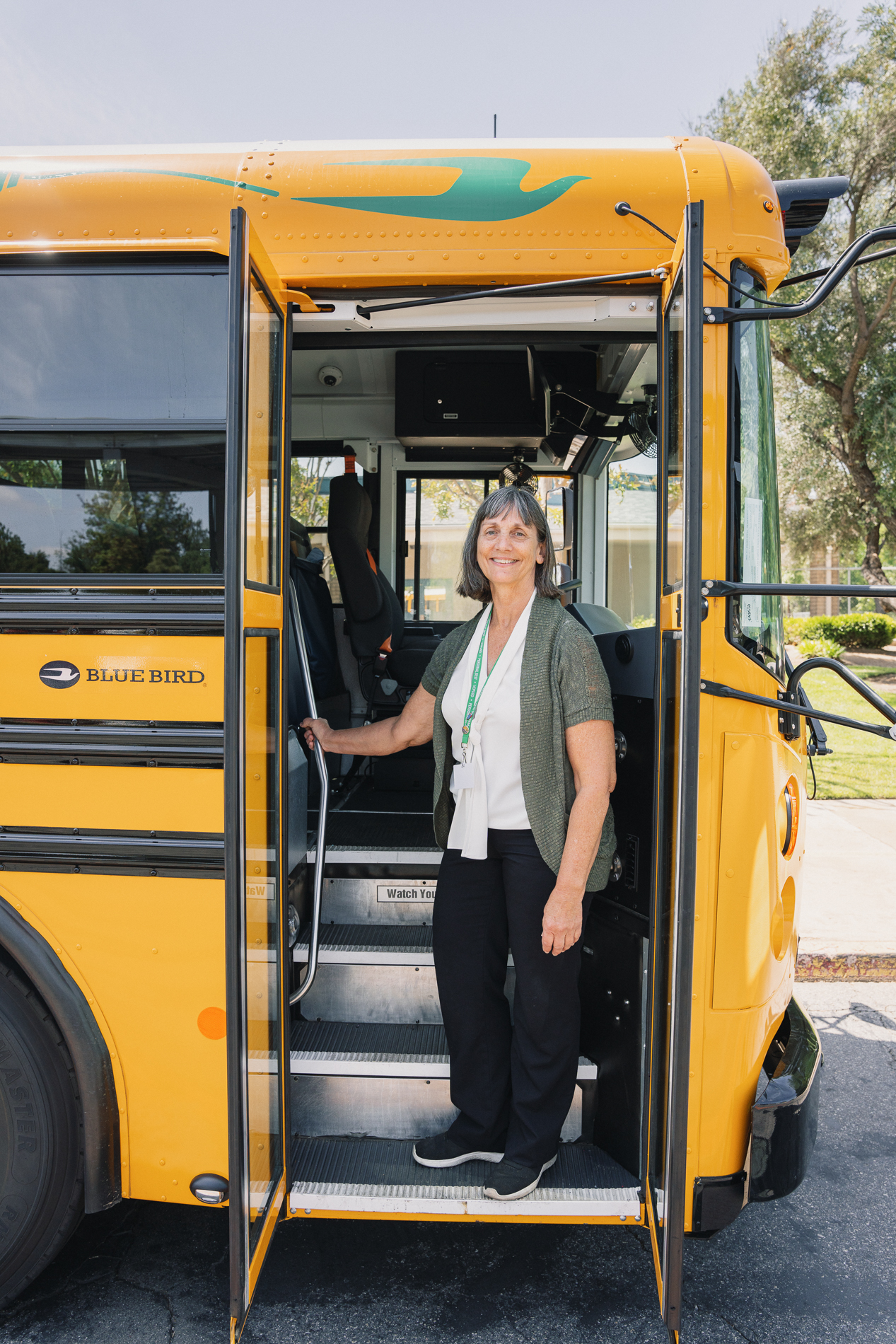 Smiling woman standing at the entrance of an open yellow Blue Bird school bus holding the handrail.