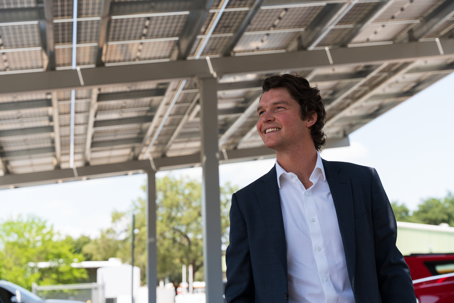 Man in a dark suit and white shirt smiling under a solar panel canopy outdoors.