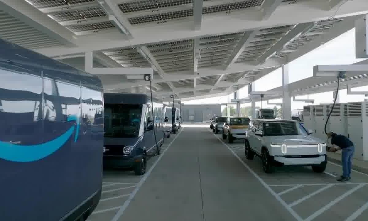 Electric delivery vans and vehicles parked at a shaded solar-powered charging station with a person plugging in one car.