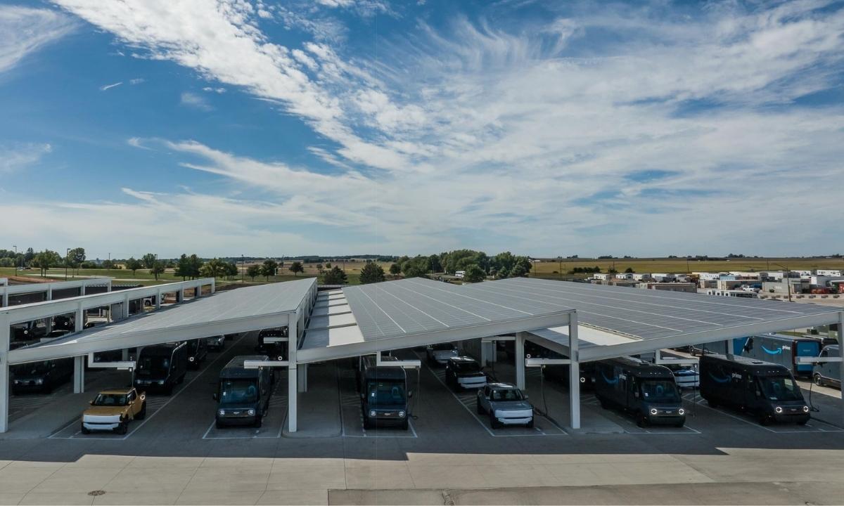 Electric delivery trucks parked under large solar panel canopies in an outdoor lot under a blue sky.