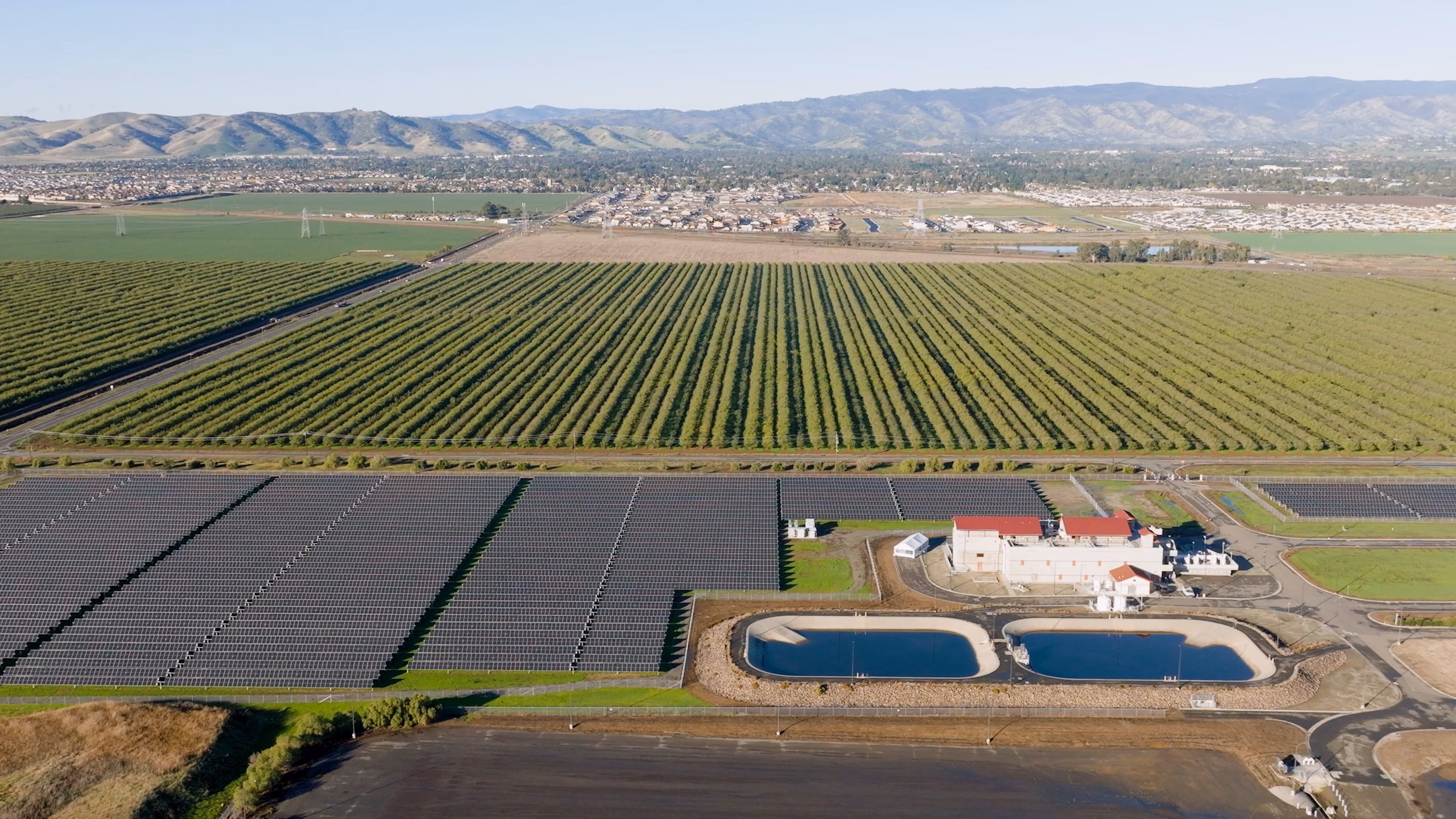 Aerial view of a solar panel farm next to orchards and two water treatment ponds with mountains in the background.