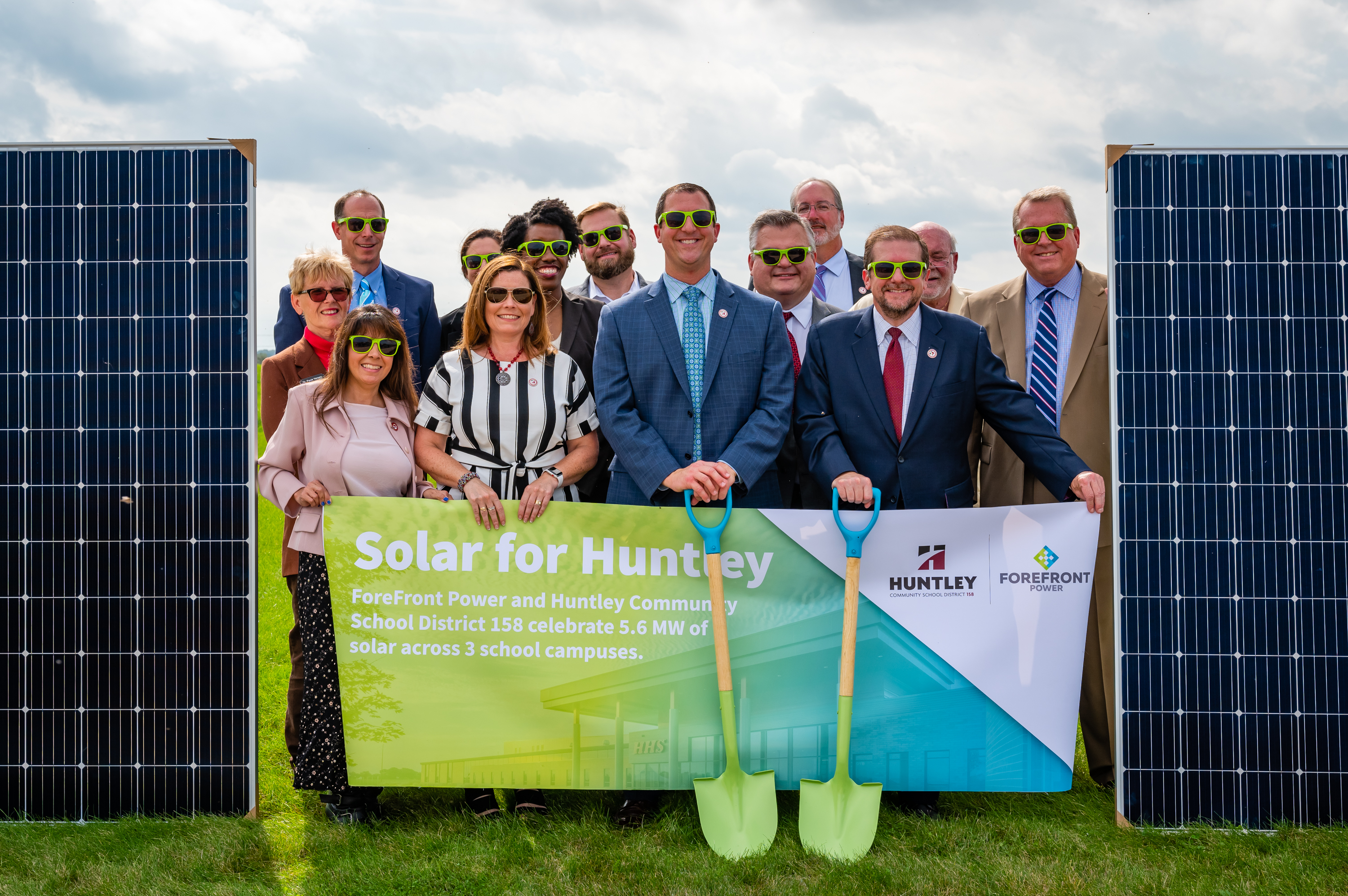 Group of people wearing green sunglasses hold a banner about solar power with two green shovels on grass.