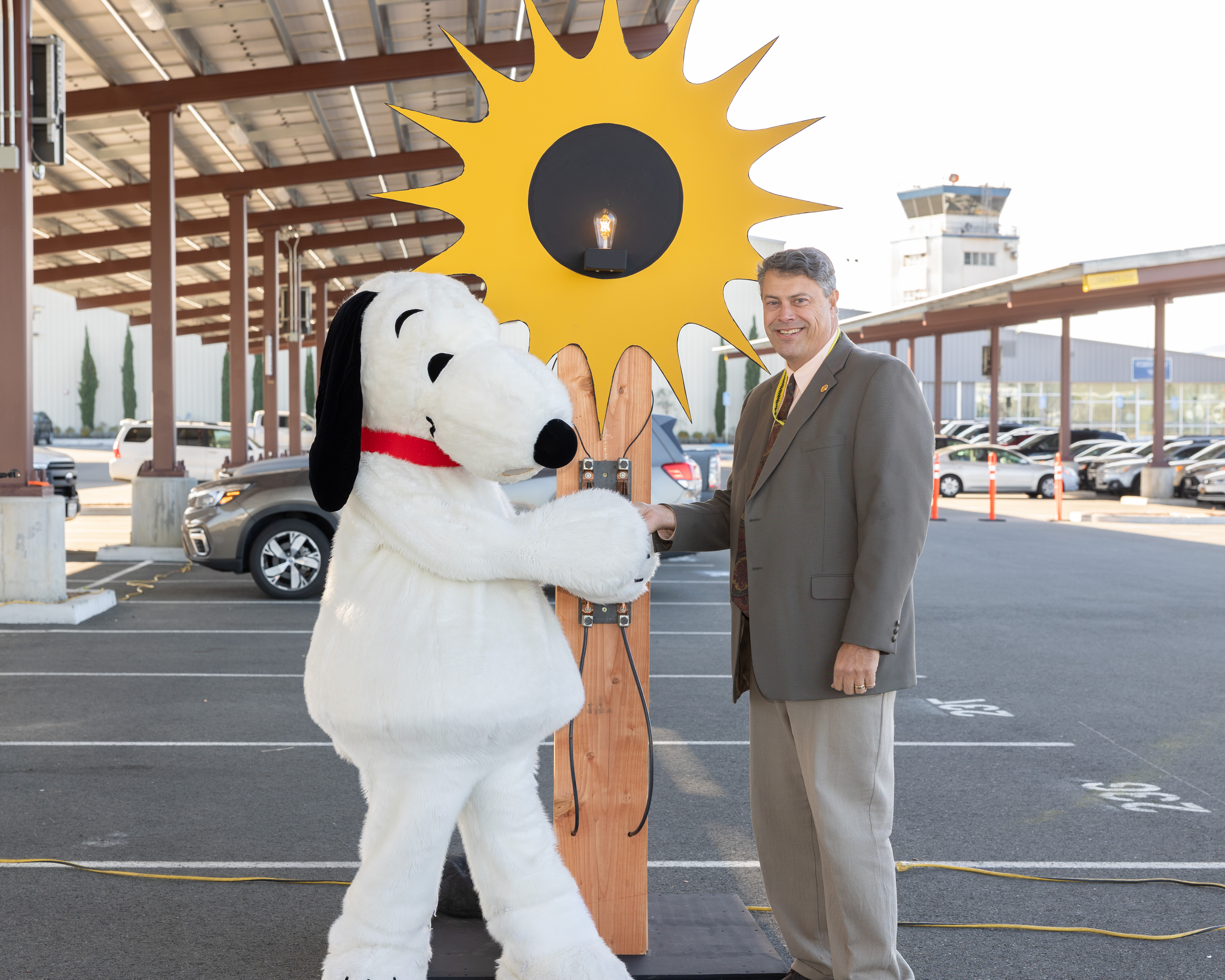 Person in Snoopy costume shaking hands with a man in a suit in a parking lot with a yellow sun-shaped light fixture.
