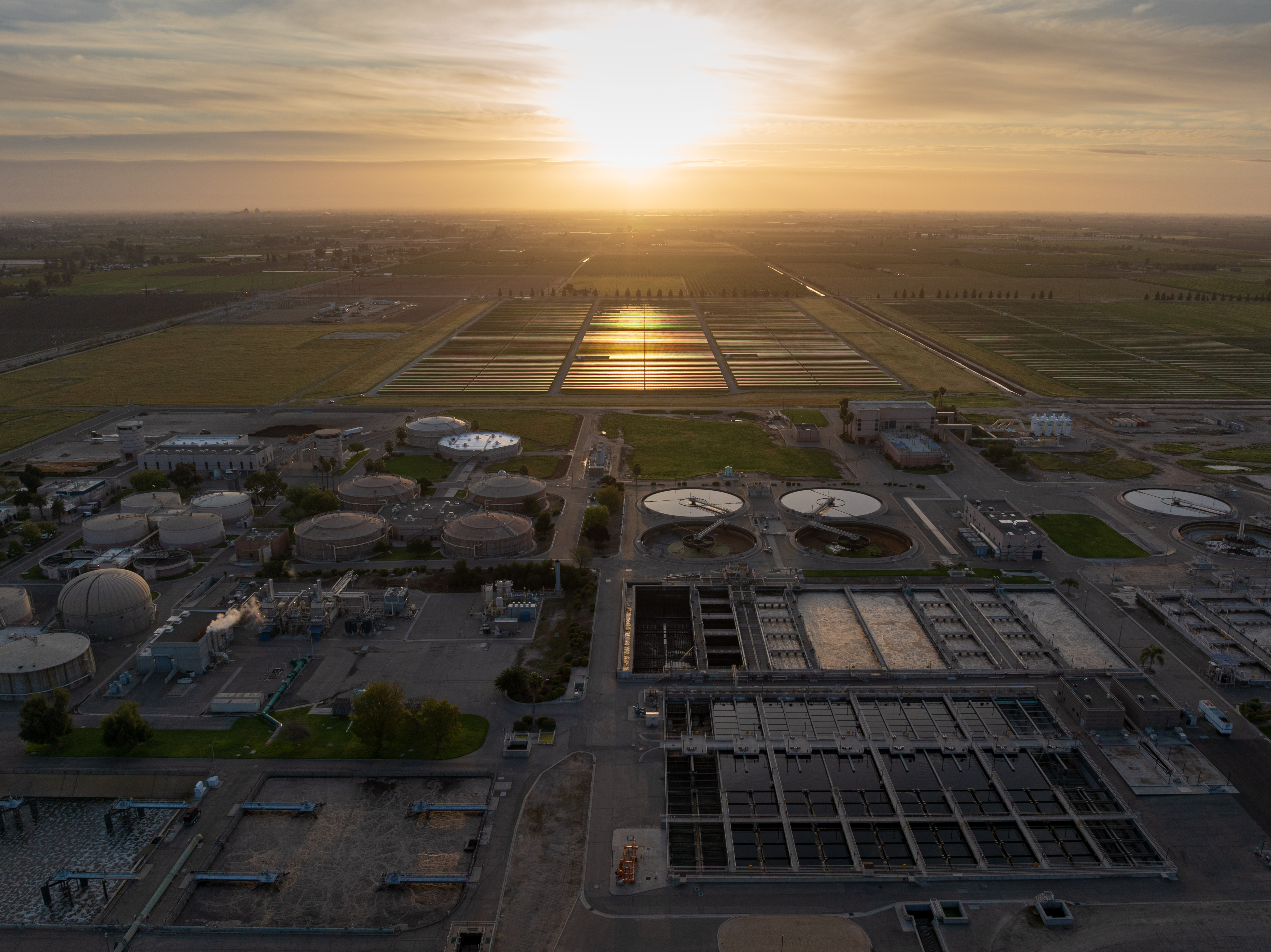 Aerial view of a water treatment facility with circular tanks and agricultural fields at sunset.