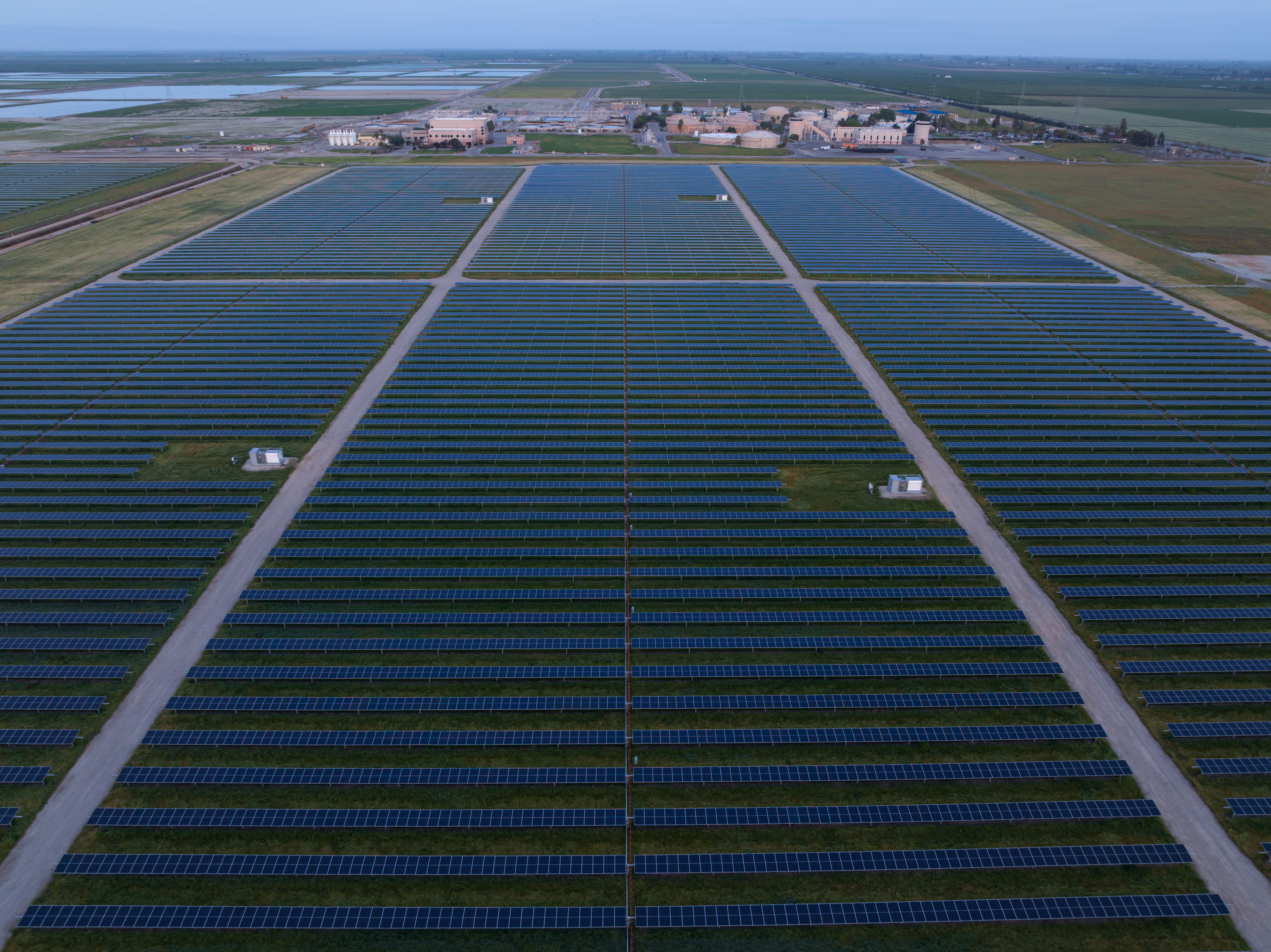 Aerial view of a large solar farm with rows of solar panels and buildings in the background.