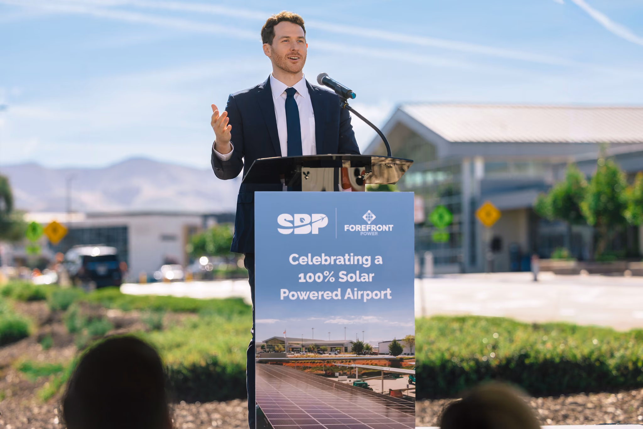 Man in suit speaking at podium celebrating a 100% solar powered airport with logos and solar panel image.
