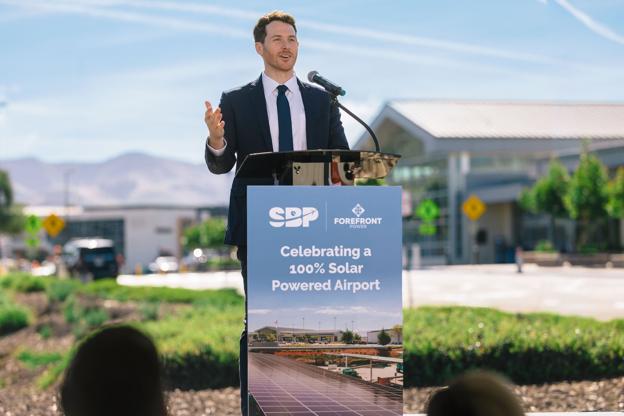 Man in suit speaking at podium celebrating a 100% solar powered airport with logos and solar panel image.
