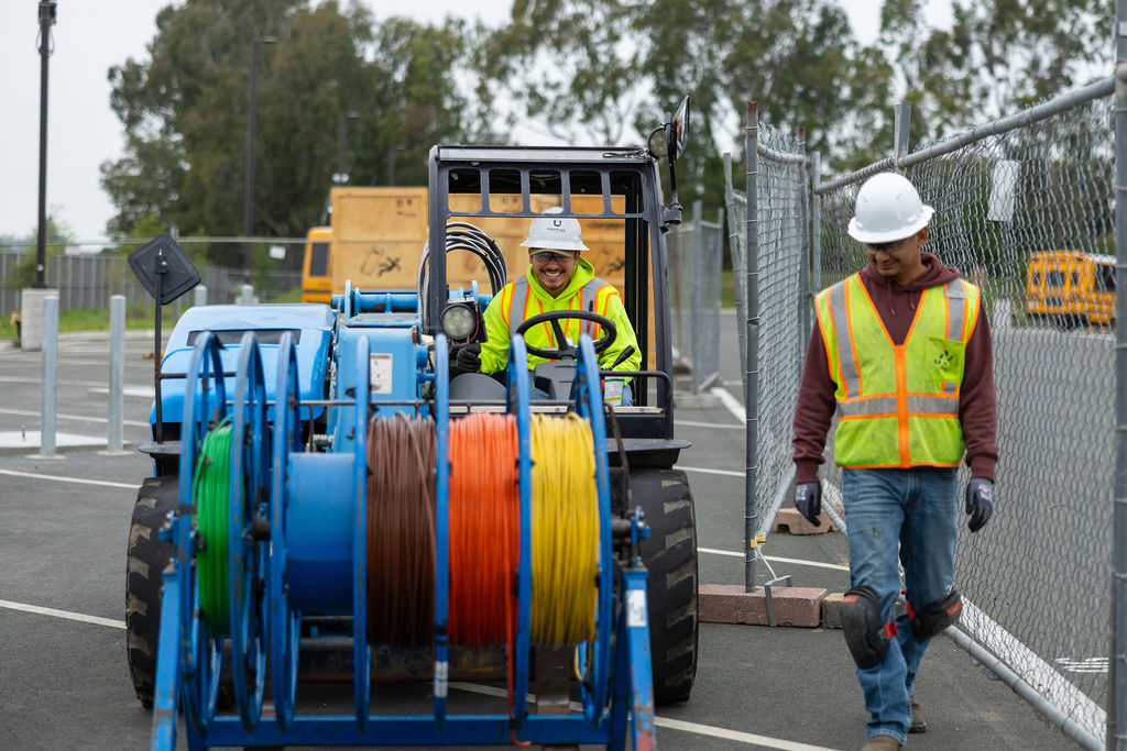 Two construction workers wearing hard hats and safety vests near cable reels and machinery.