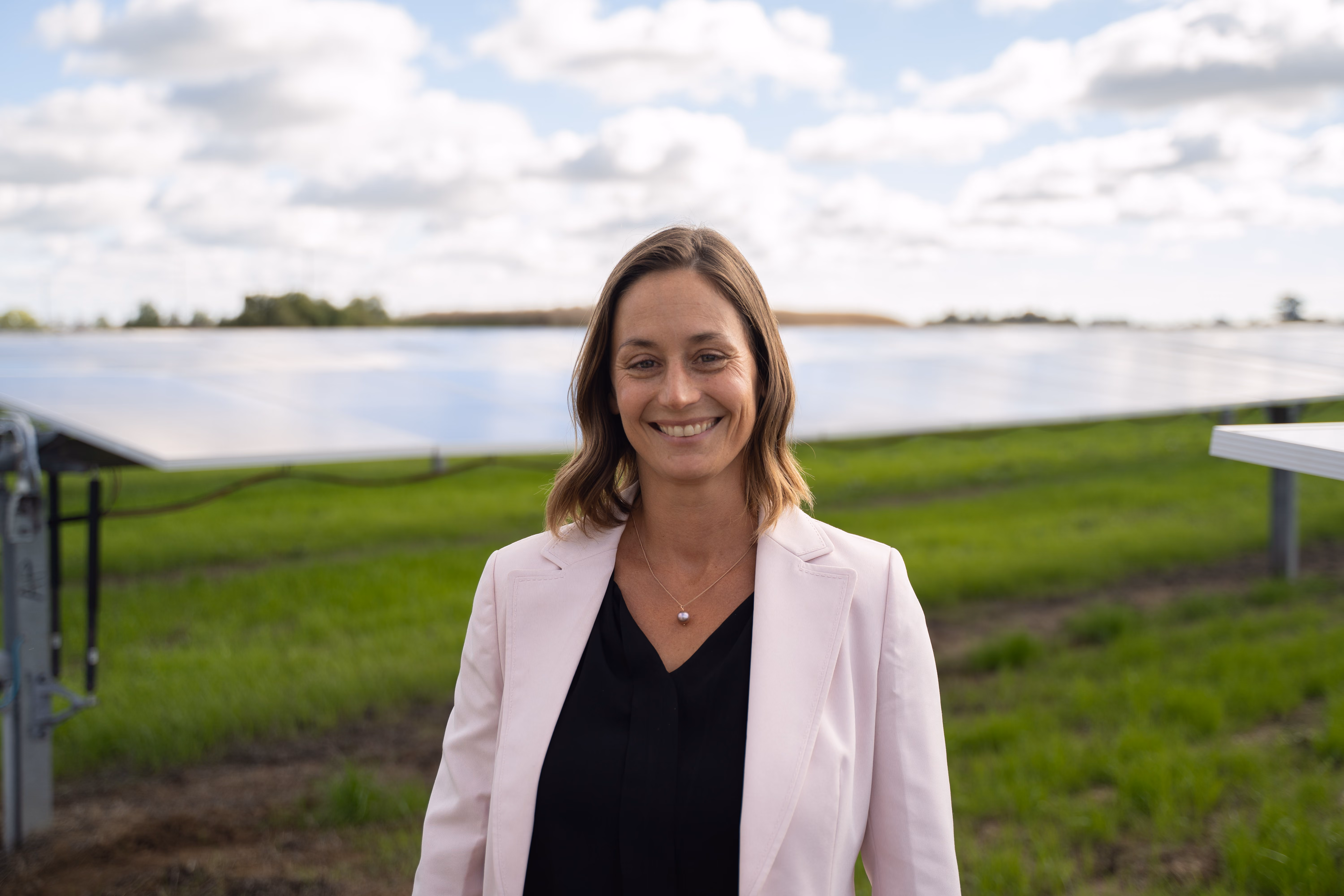 Smiling woman in light blazer standing in front of solar panels in a green field under a partly cloudy sky.