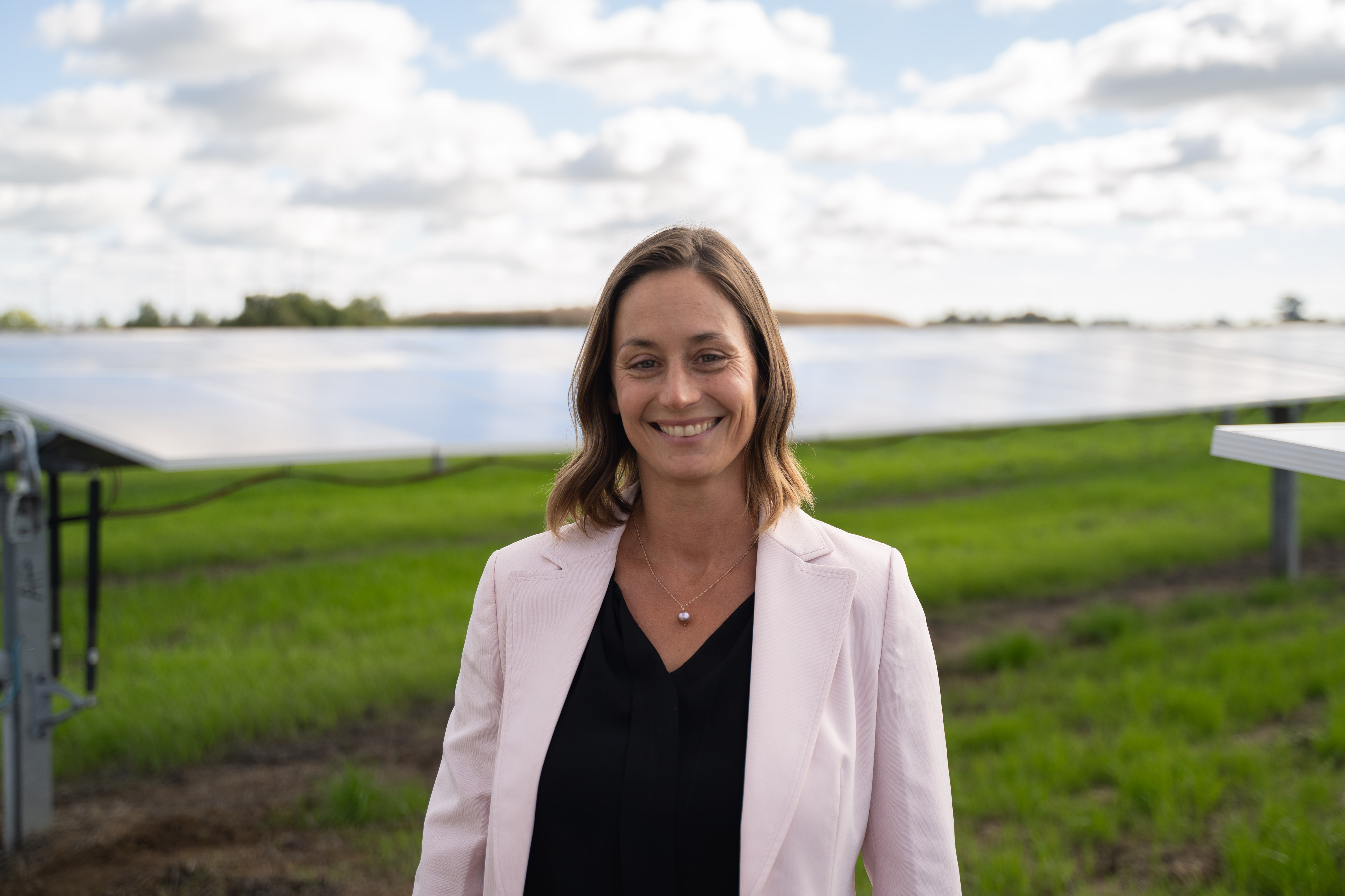 Smiling woman in light blazer standing in front of solar panels in a green field under a partly cloudy sky.