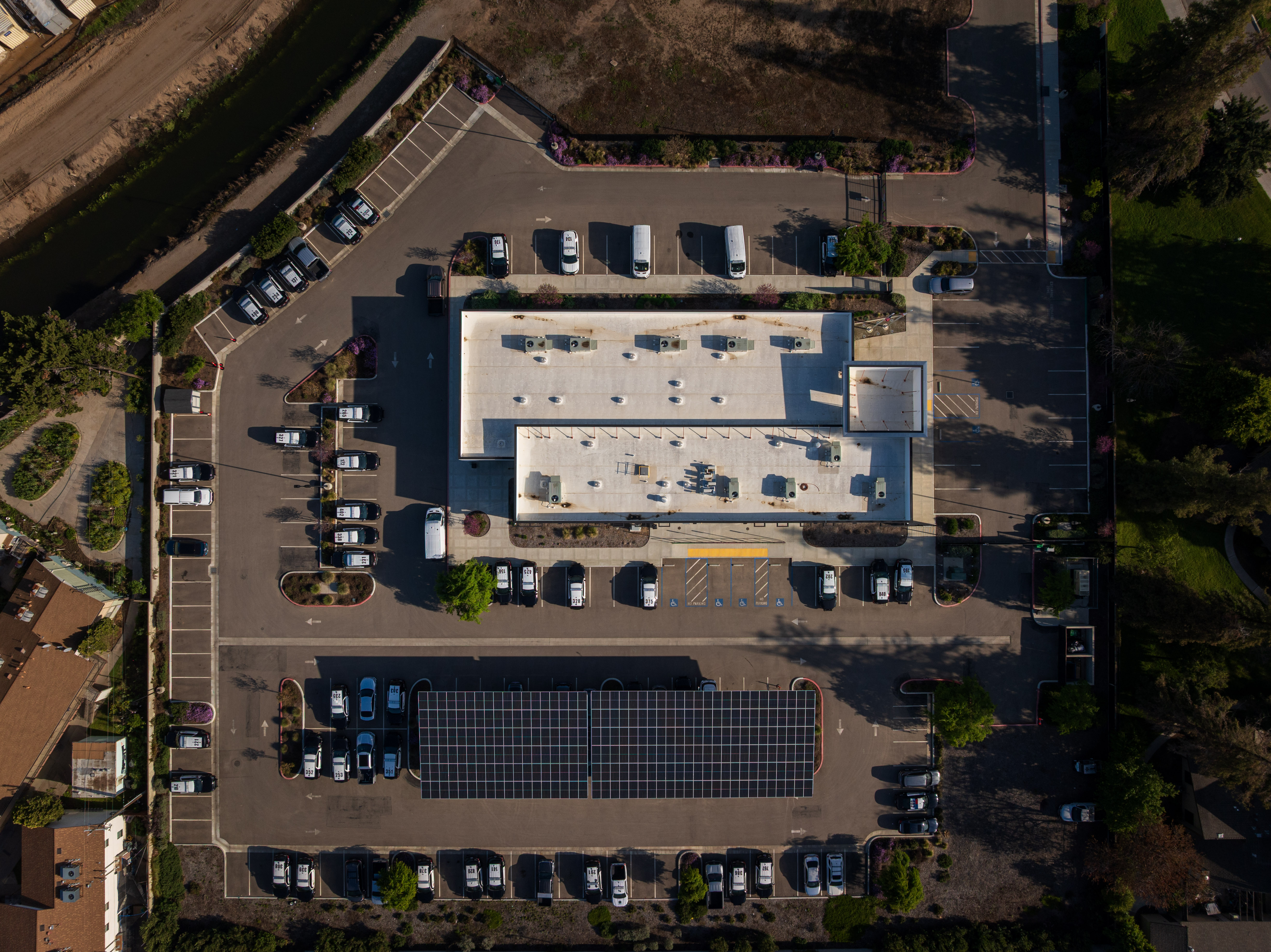 Aerial view of a large building with white roof and parked cars around a solar panel-covered parking area.