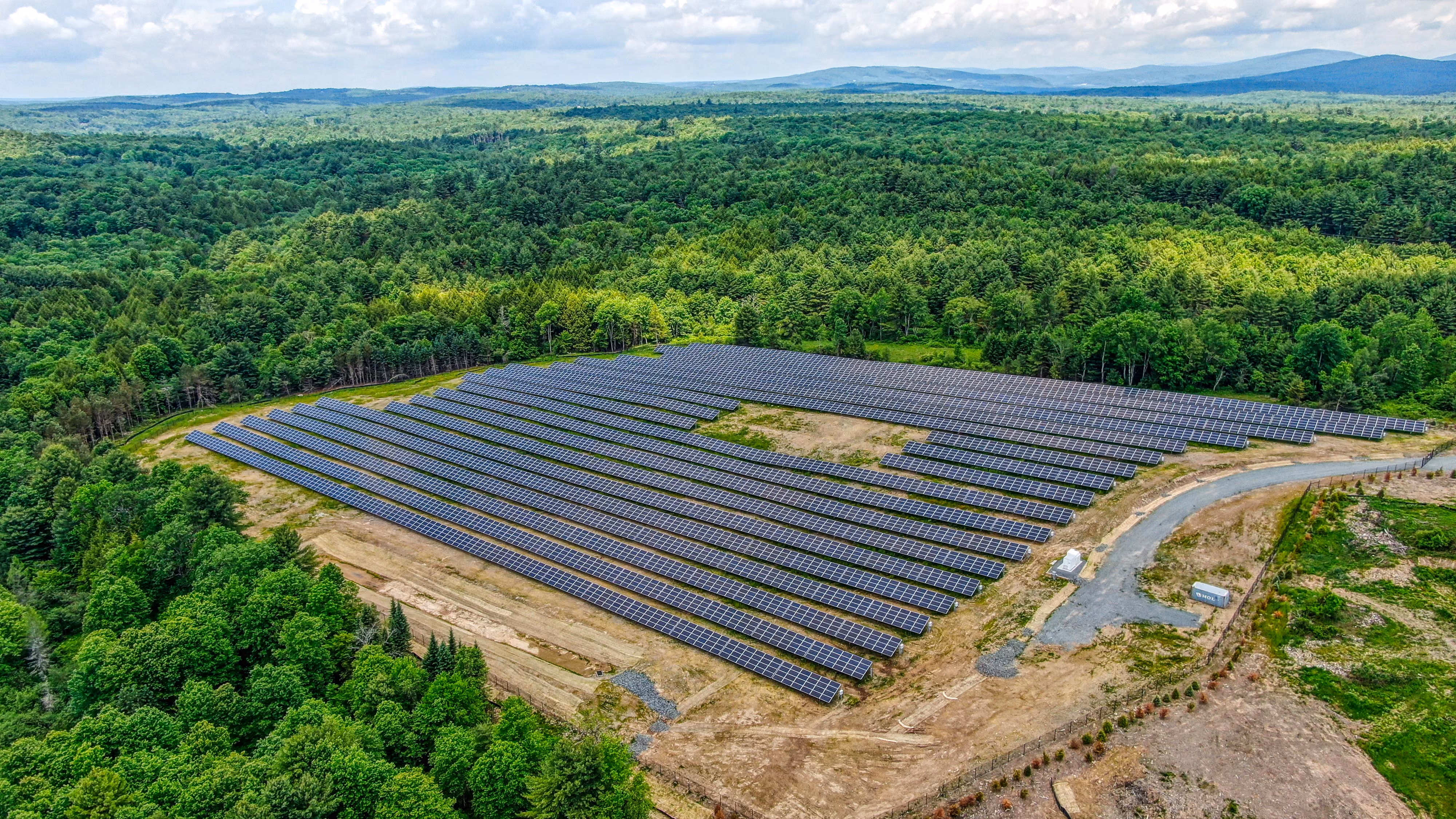 Large solar panel array on cleared land surrounded by dense green forest under a partly cloudy sky.