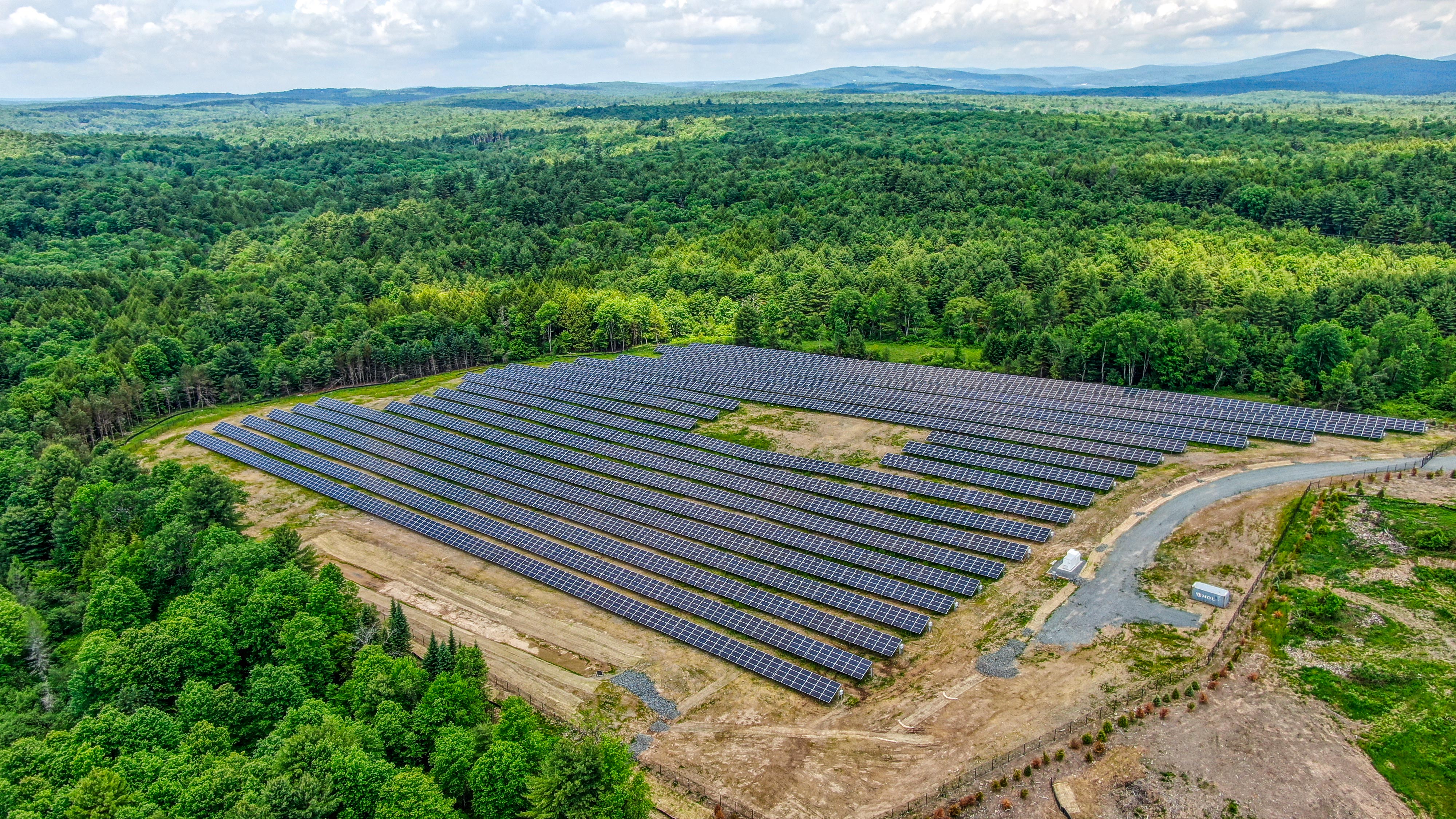 Large solar panel array on cleared land surrounded by dense green forest under a partly cloudy sky.