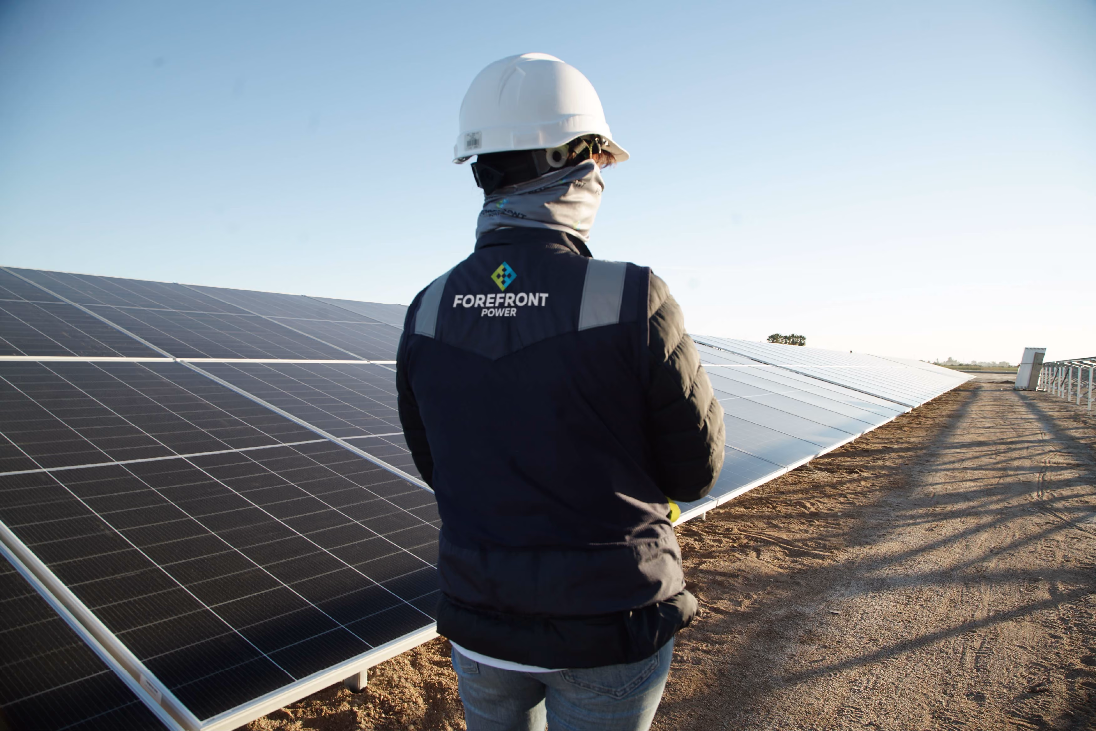 Person wearing a Forefront Power vest and hard hat inspecting solar panels outdoors.