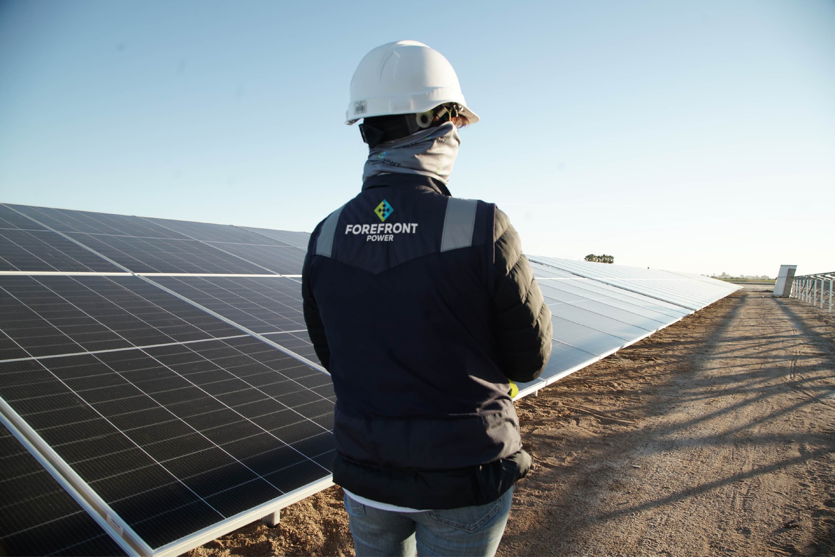 Person wearing a Forefront Power vest and hard hat inspecting solar panels outdoors.