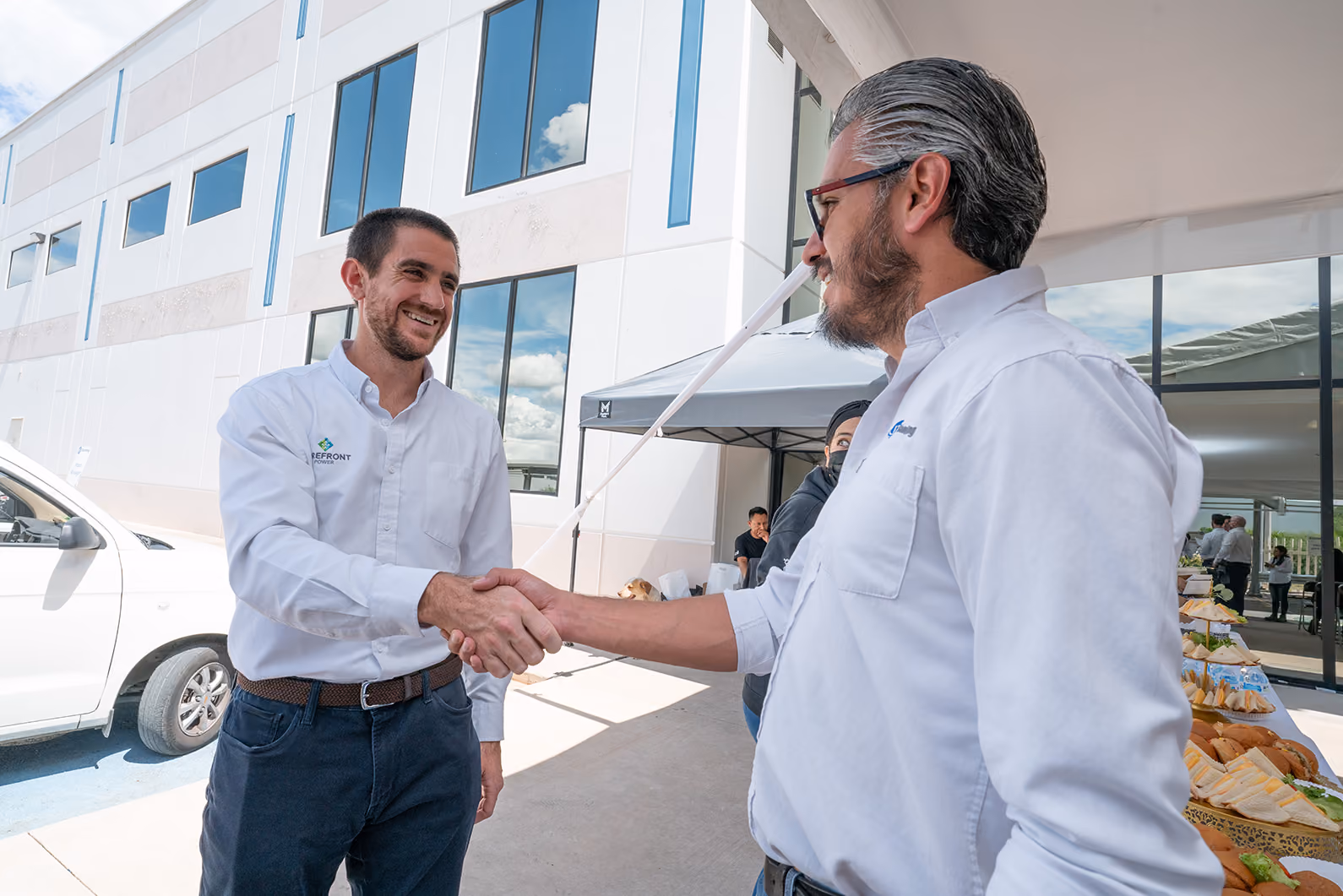 Two men in white shirts shaking hands outdoors next to a building and food table.