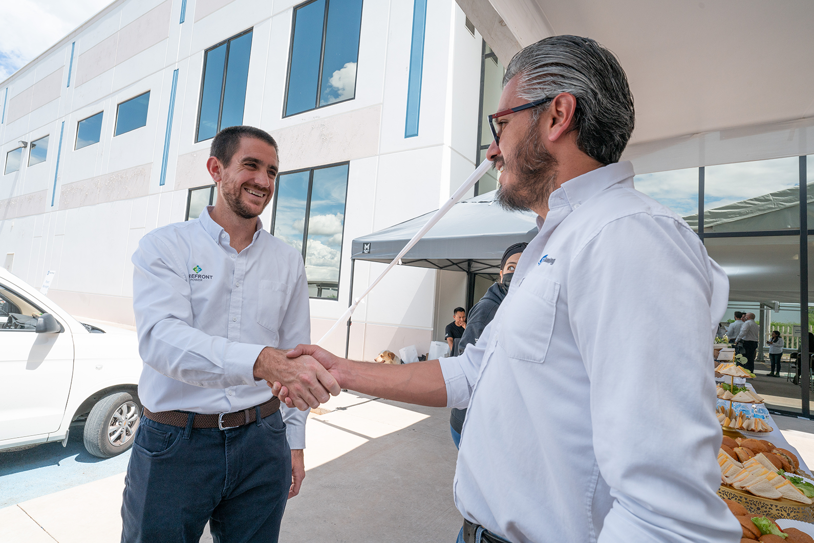 Two men in white shirts shaking hands outdoors next to a building and food table.