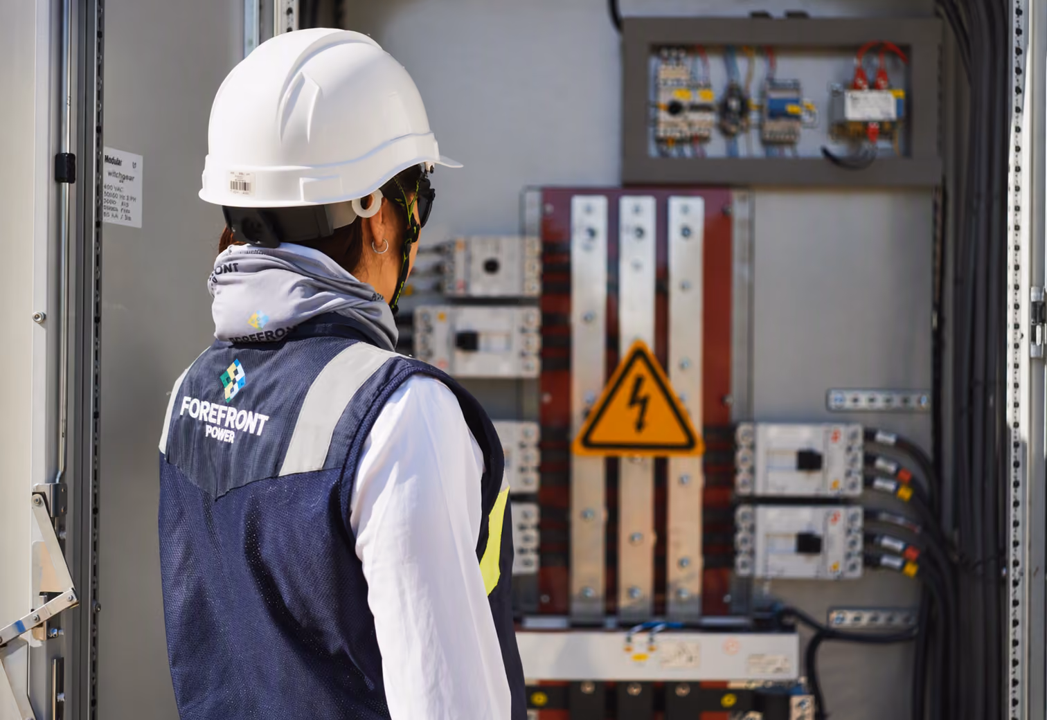 Electrician wearing a white hard hat and Forefront Power vest inspects an electrical panel with a hazard sign.