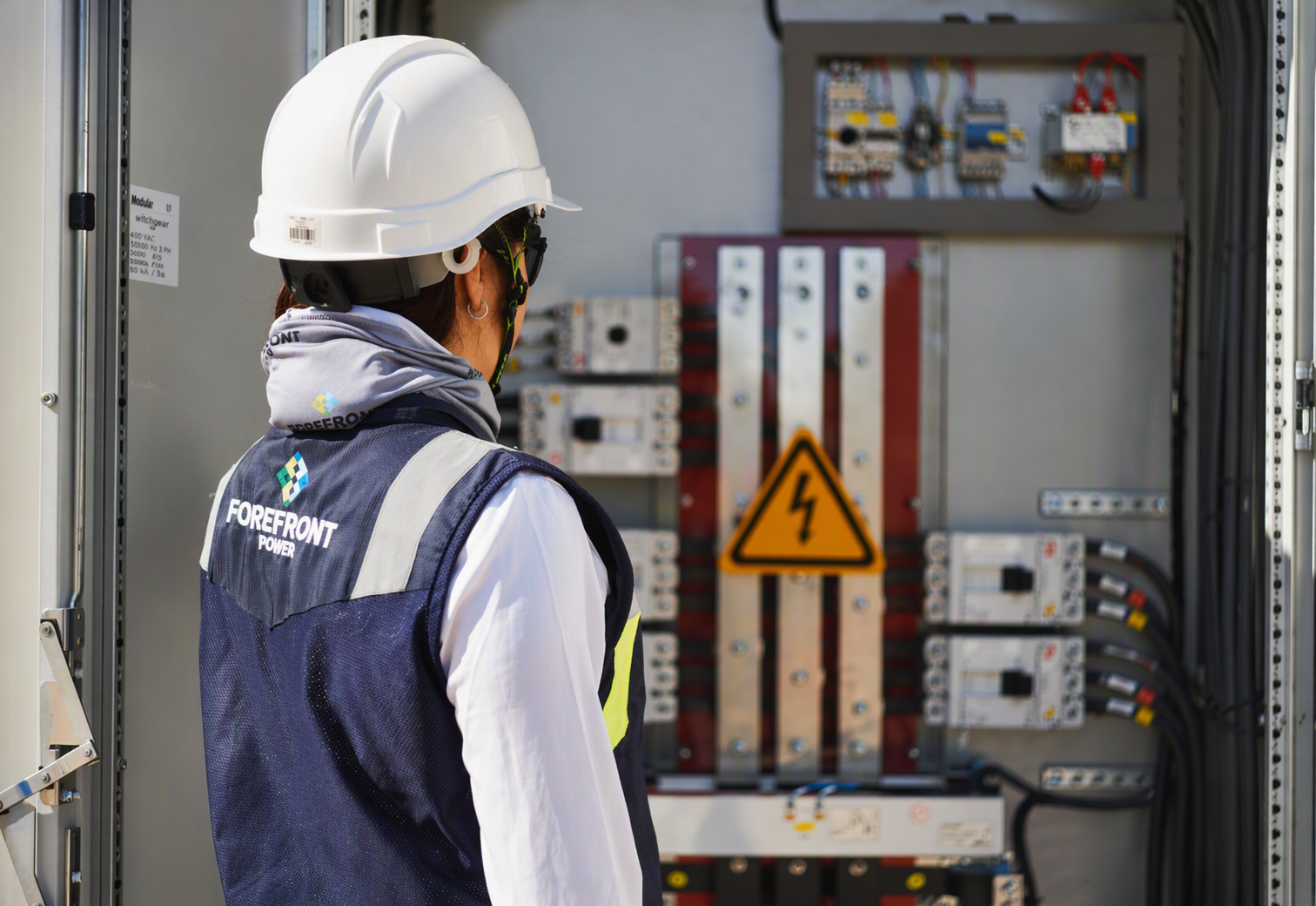 Electrician wearing a white hard hat and Forefront Power vest inspects an electrical panel with a hazard sign.