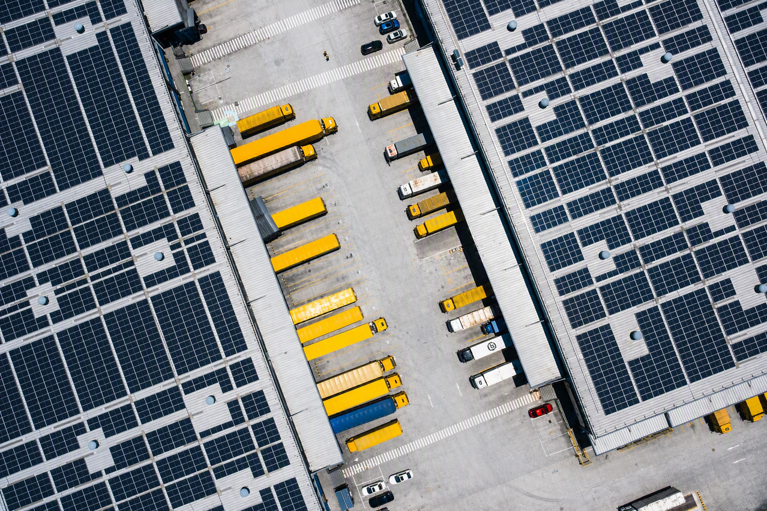Aerial view of warehouse rooftops with solar panels and multiple yellow trucks parked below.