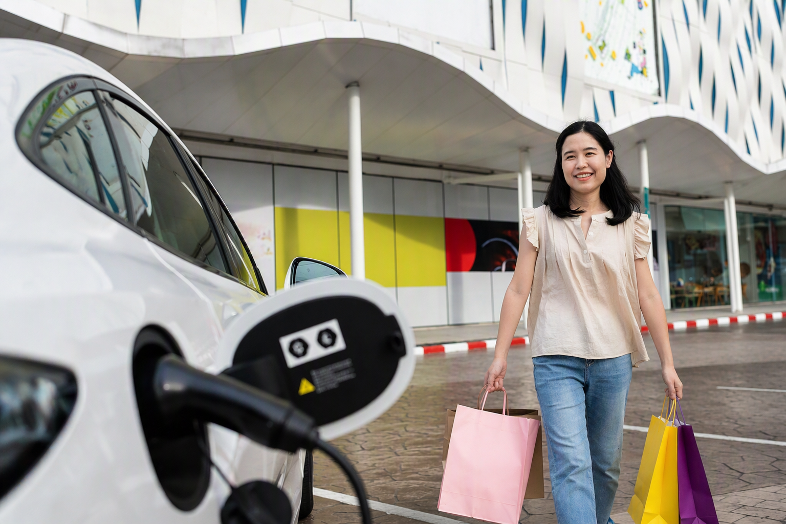 Woman with shopping bags walking past an electric car charging outside a modern building.