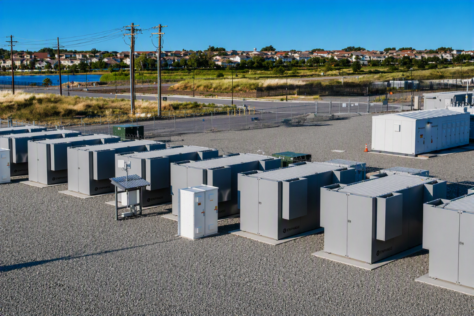 Row of large gray electrical battery storage units on gravel near a residential area and power lines.