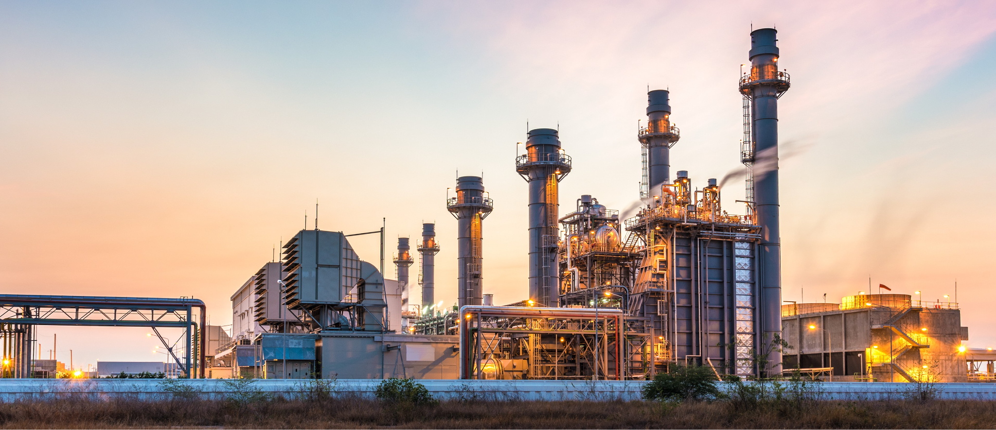 Industrial power plant with tall smokestacks and equipment at sunset under a clear sky.
