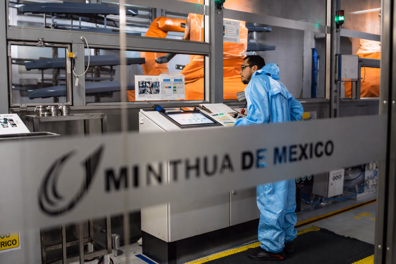 Man in blue protective suit operating control panel in a factory with robotic arms in the background.