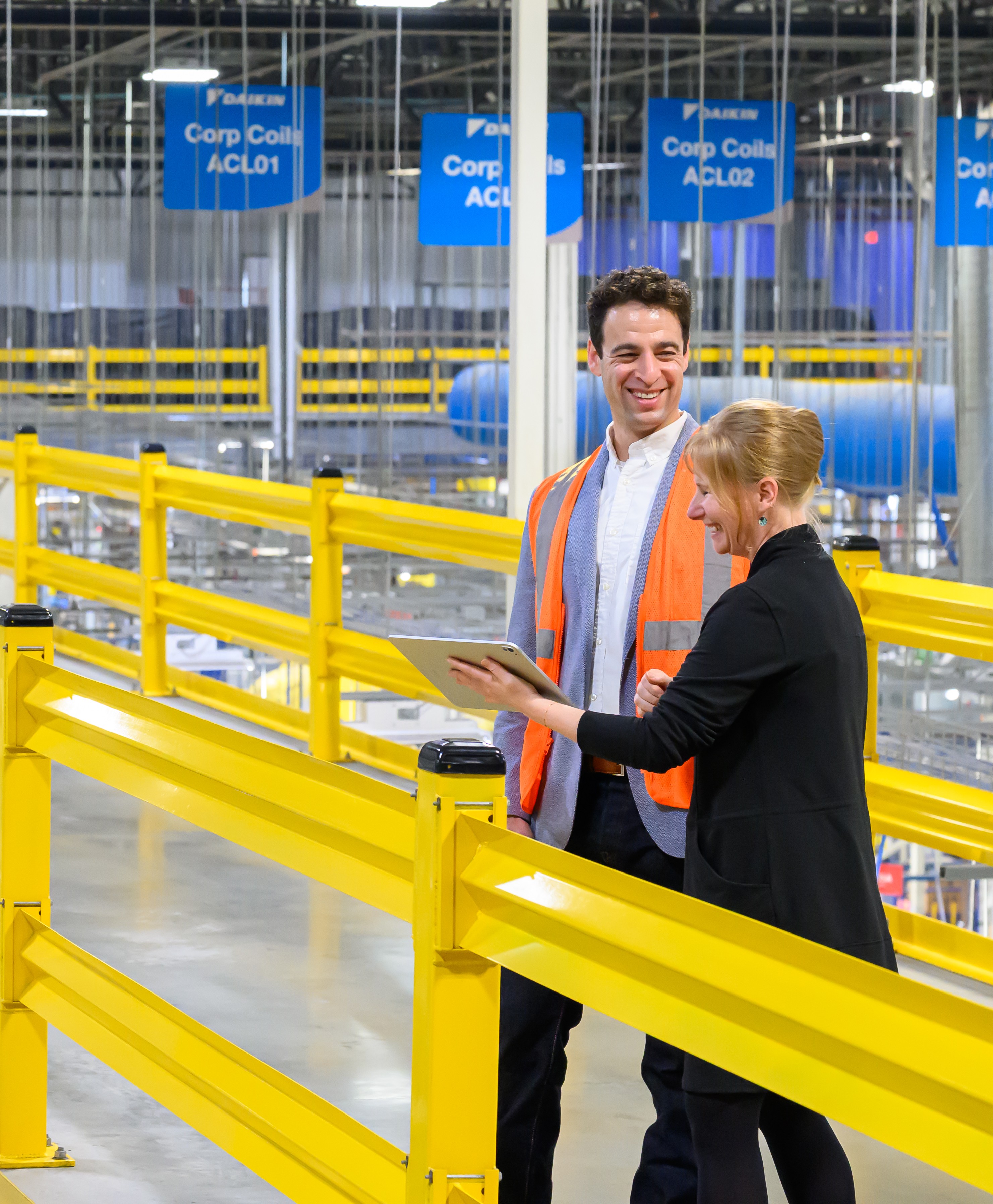 Two factory workers with a tablet standing inside a warehouse with yellow safety railings and blue signs.