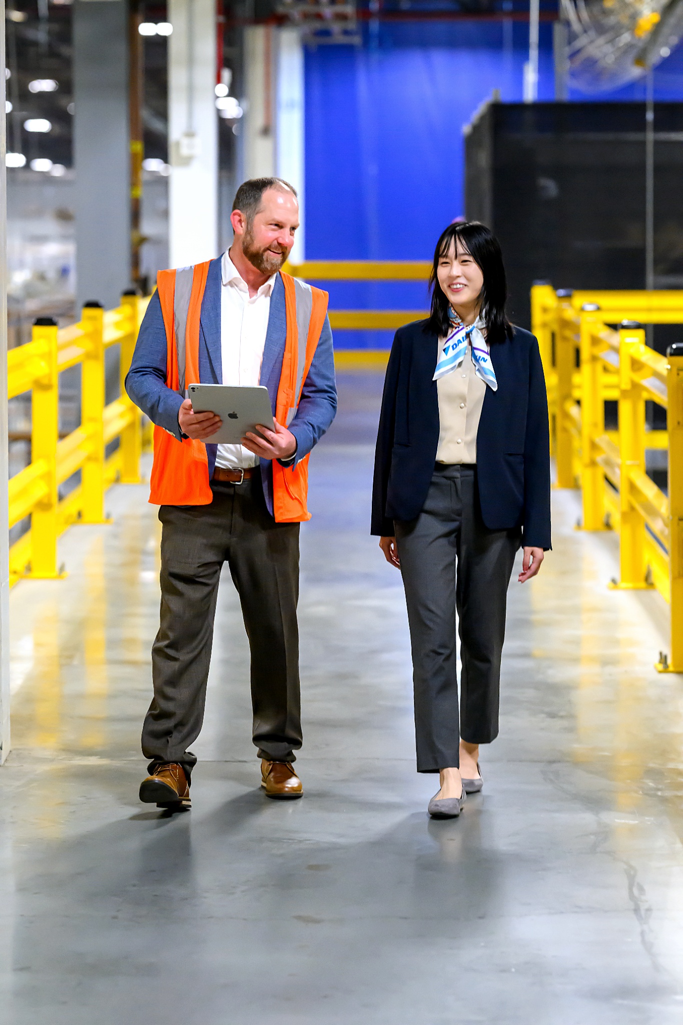 Man in orange safety vest holding tablet and woman in business attire walking in warehouse aisle.