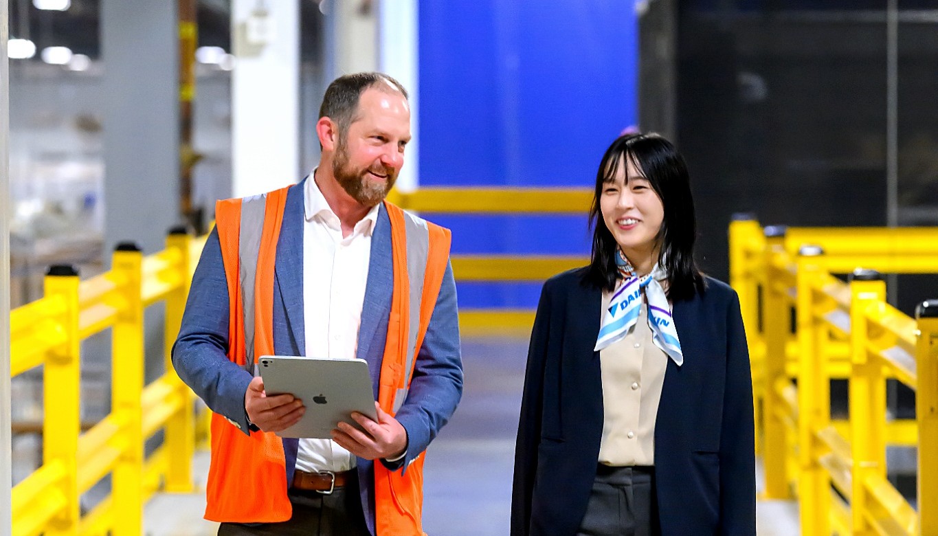 Man in orange safety vest holding tablet walks and talks with smiling woman in business attire in industrial setting.