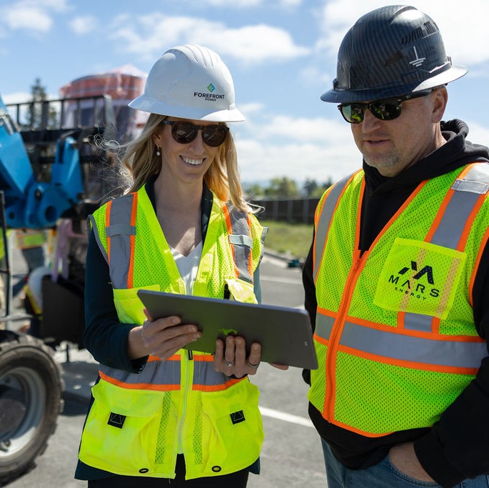 Two construction workers wearing hard hats and reflective vests looking at a tablet outdoors.