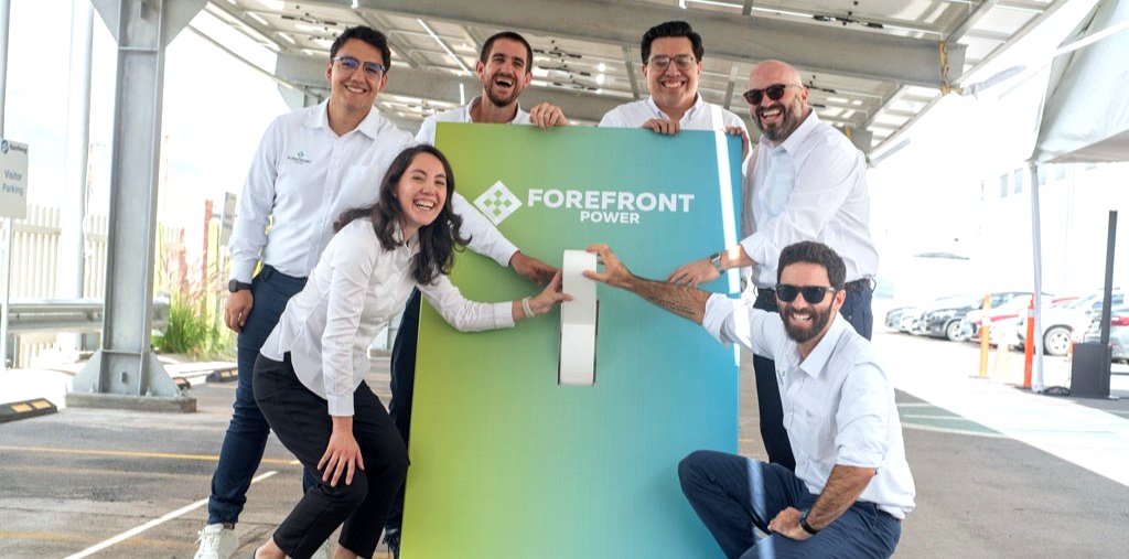 Group of six people in white shirts smiling around a large Forefront Power switch outdoors.