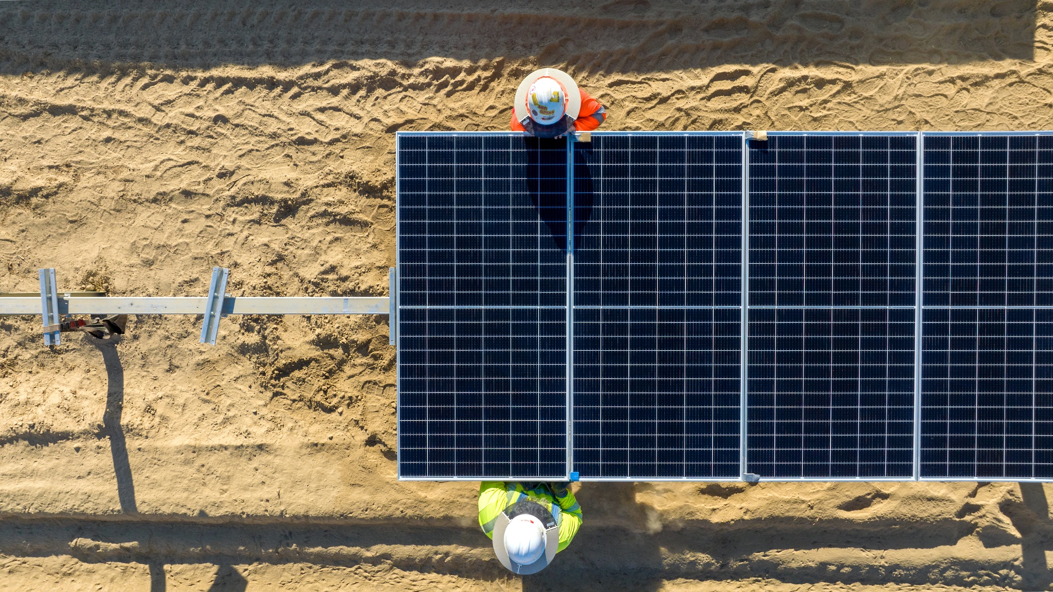 Overhead view of two workers installing large solar panels on sandy ground.
