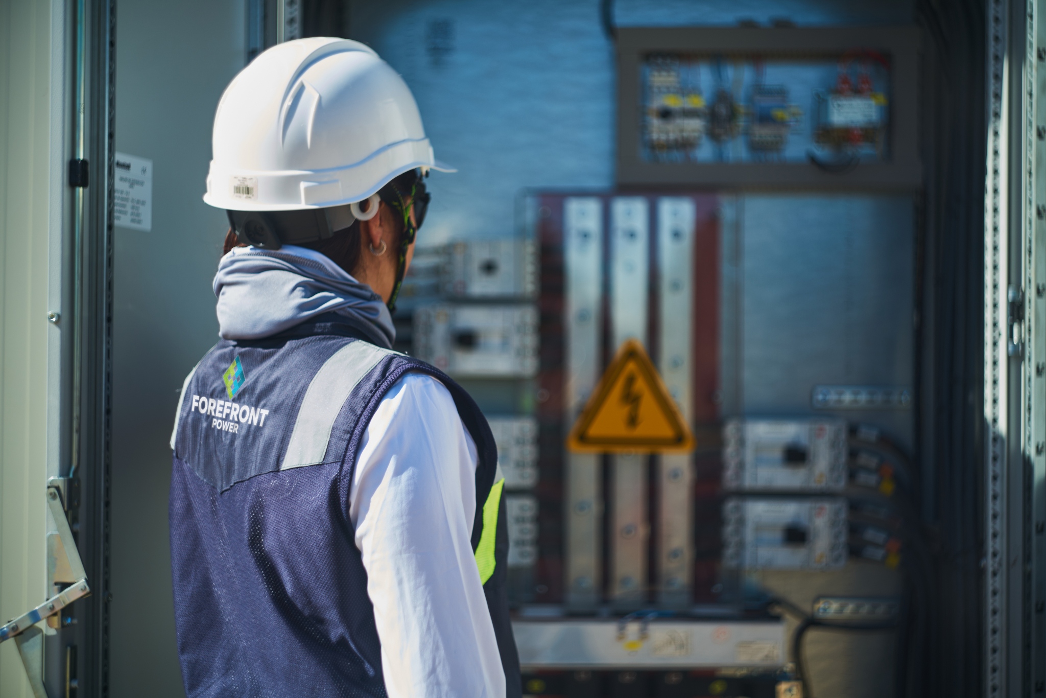 Worker in a white helmet and Forefront Power vest inspecting electrical panel with warning sign.