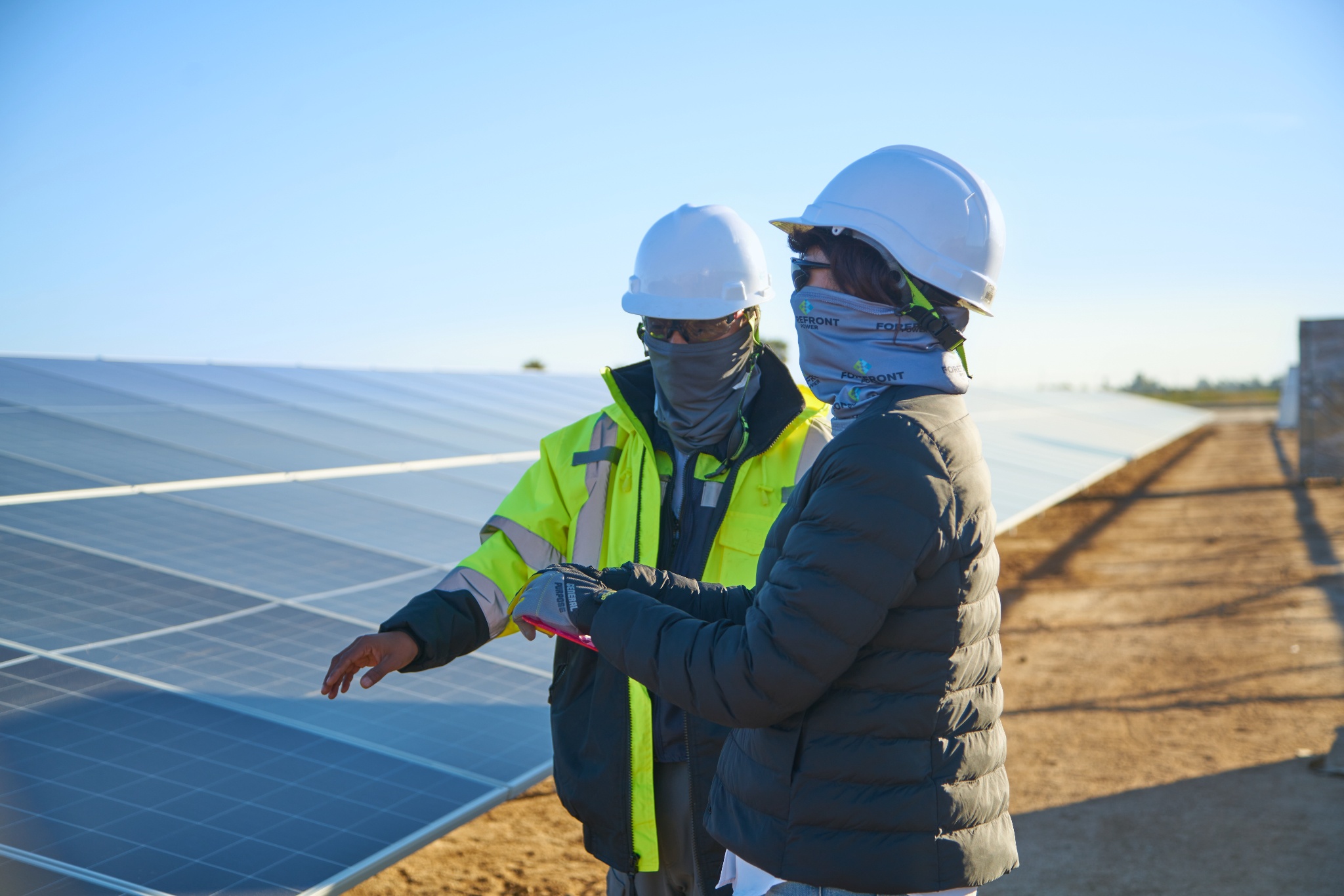 Two workers wearing helmets and jackets inspecting solar panels outdoors on a sunny day.
